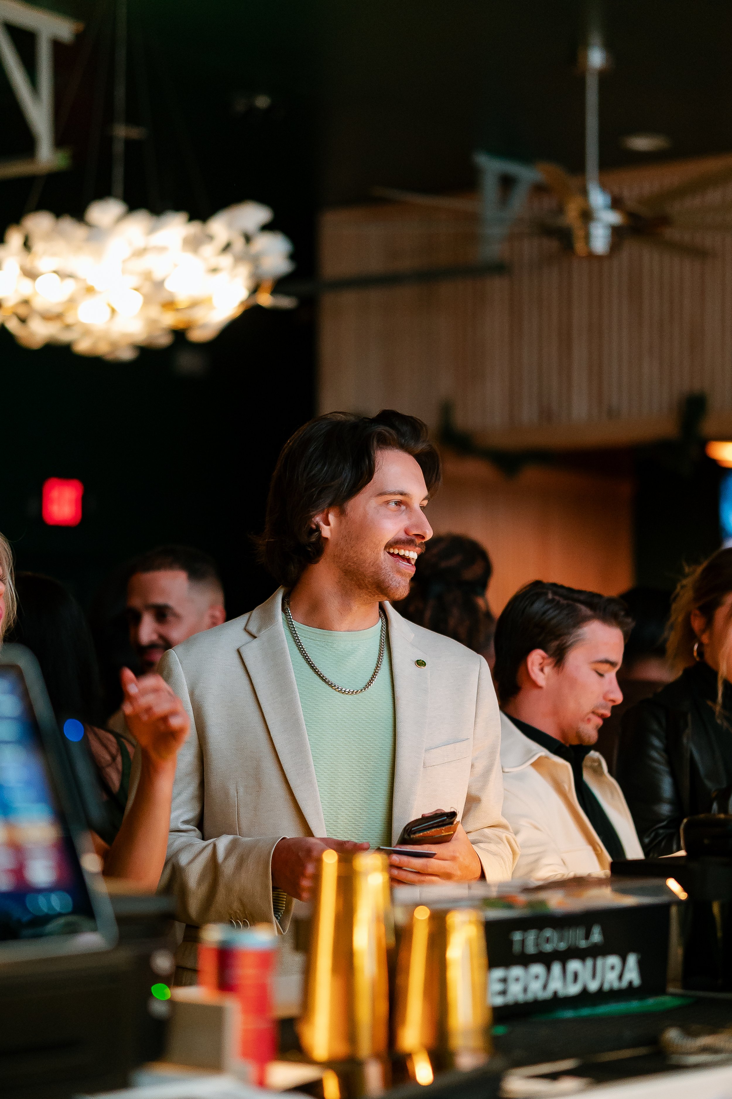 Young man smiling in a bar with others around, wearing a light blazer and holding a phone; tequila bottles visible in foreground.