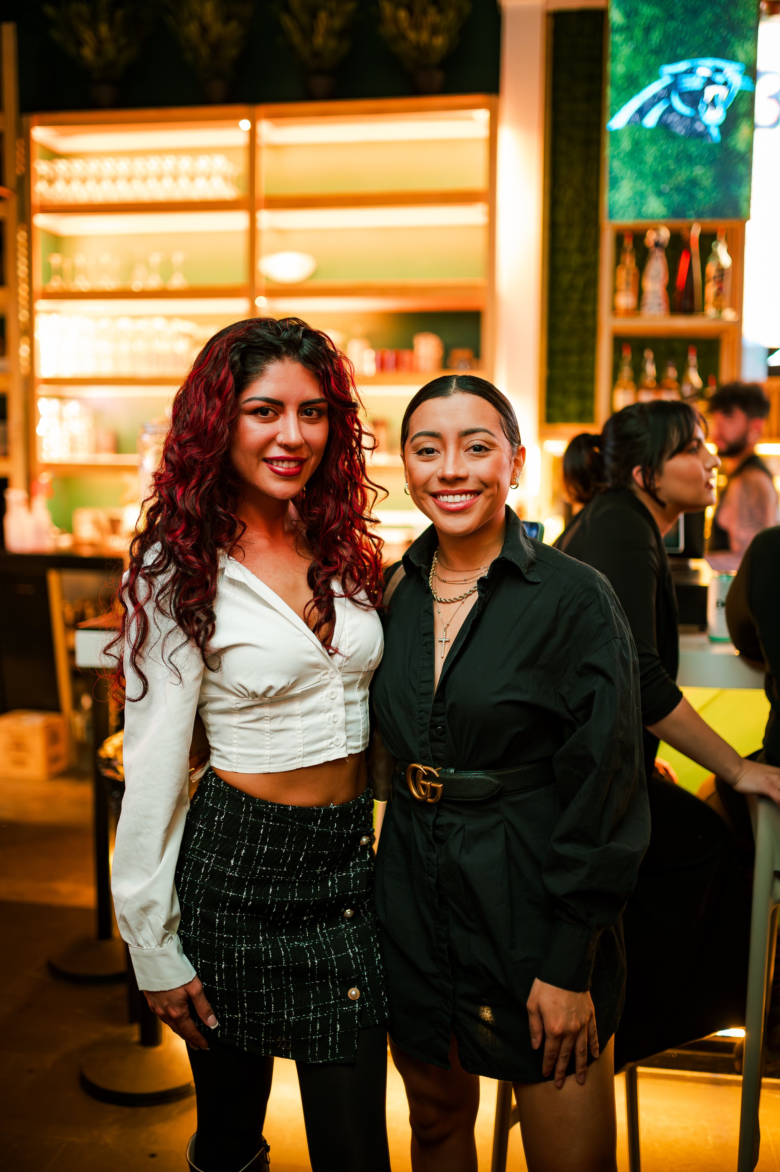 Two women posing and smiling in a bar, with shelves of bottles and glasses in the background.