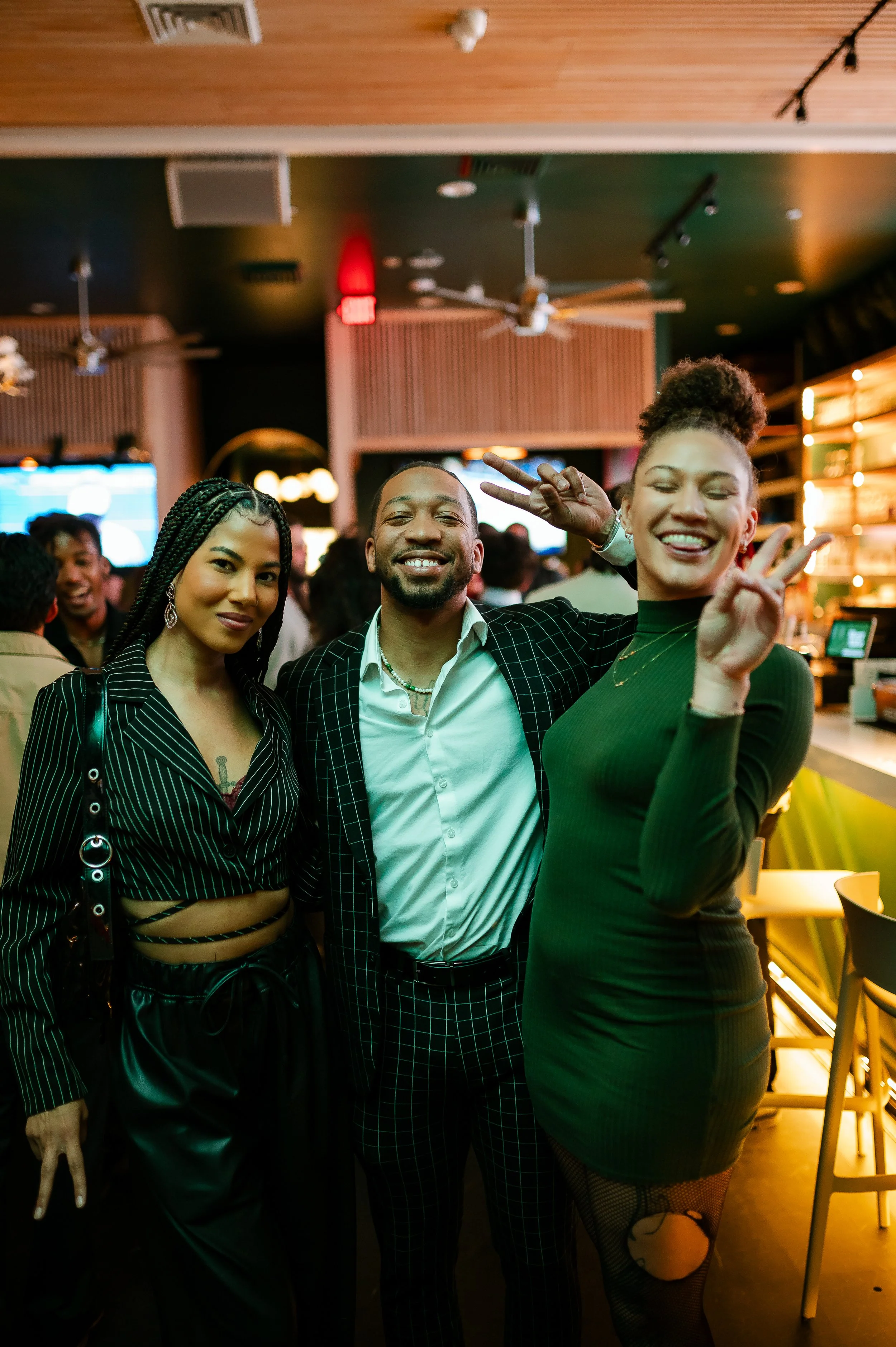 Three people smiling and posing for a photo in a lively indoor setting with modern decor. One person is wearing a pinstripe suit, another in a green dress, and the third in a stylish outfit. They appear to be enjoying a social event or party.