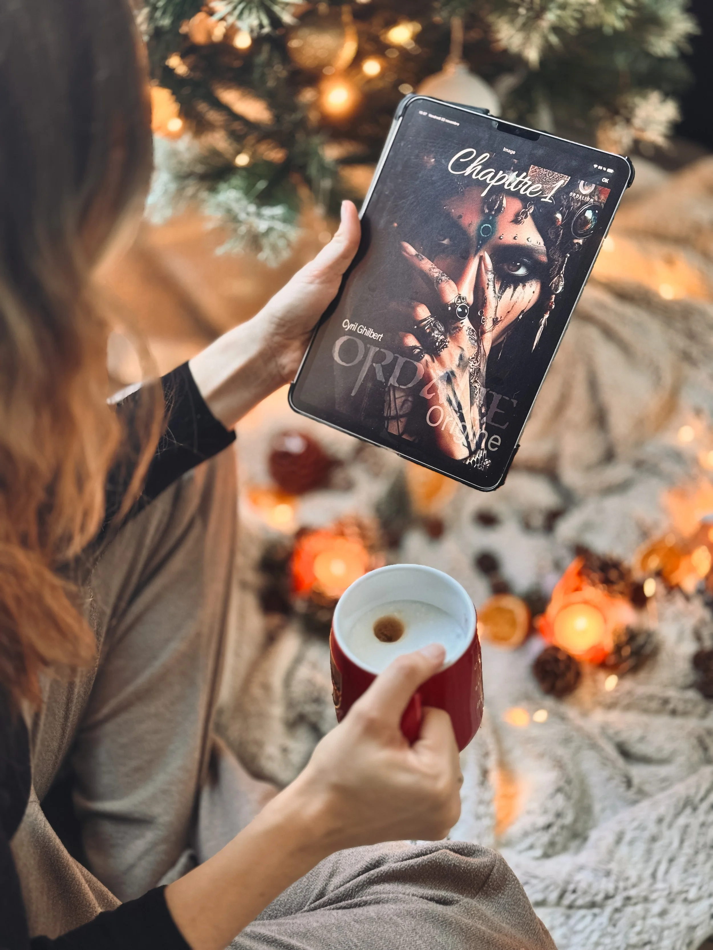 Une personne tient une tablette montrant la couverture du livre 'Ordinate' de Cym Ghilbert, devant un sapin de Noël décoré, une tasse de café ou de chocolat chaud dans l'autre main, avec des décorations de Noël par terre.