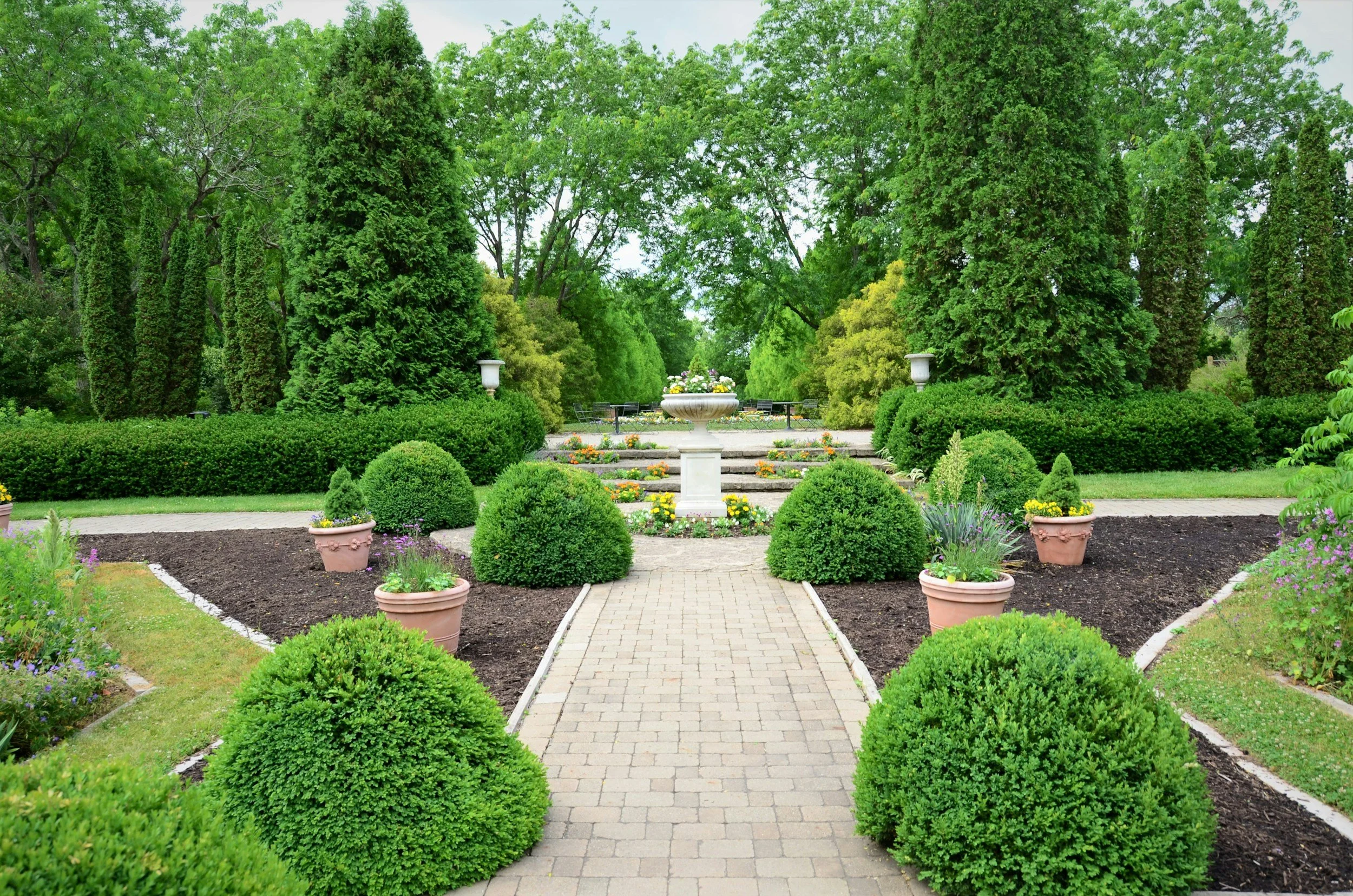 A landscaped garden with a brick pathway, green shrubs, flower pots with blooming flowers, and tall trees in the background.