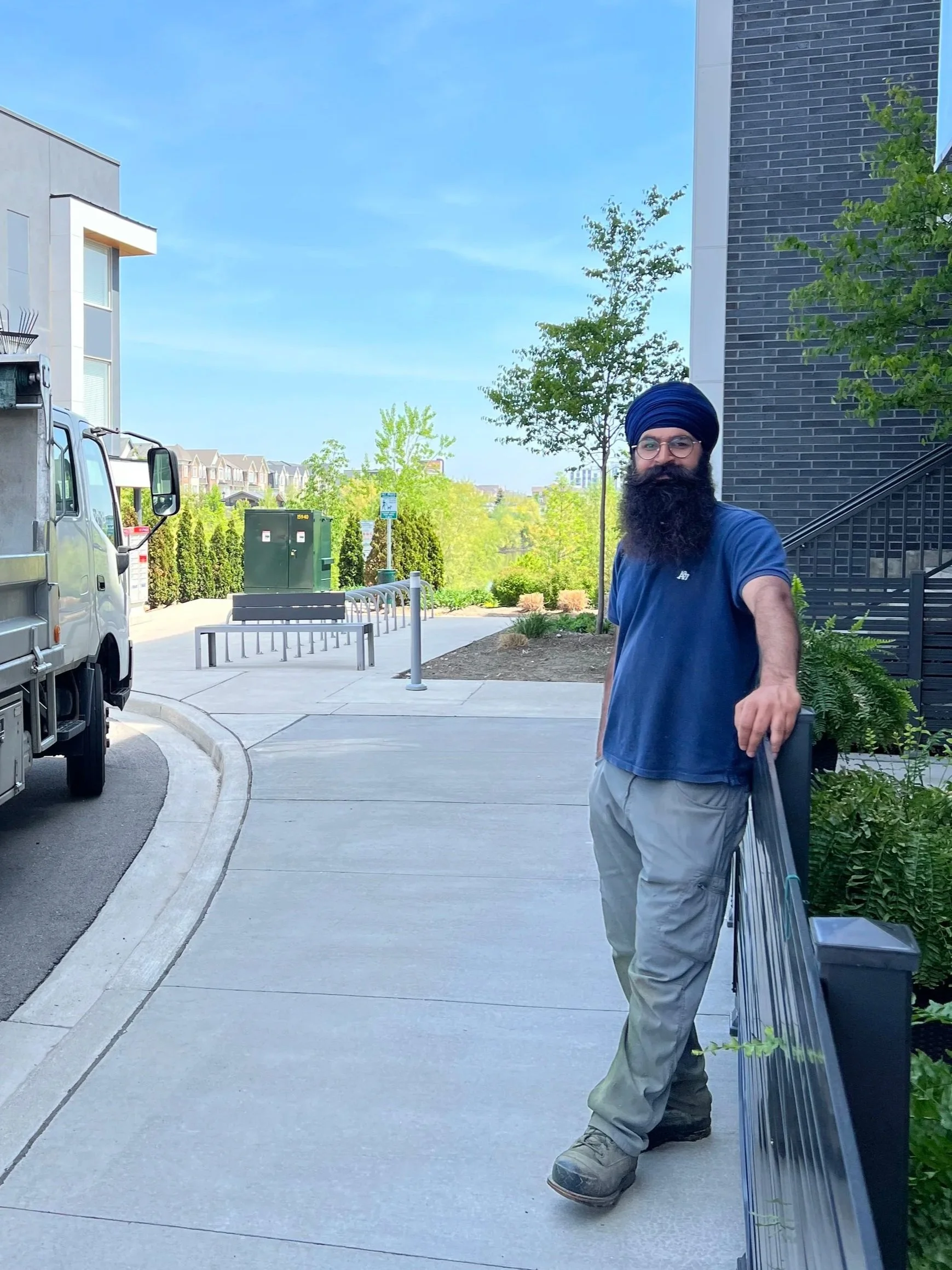 A man with a dark beard and glasses wearing a blue turban, blue shirt, and beige pants standing outside on a sidewalk, leaning on a railing next to a black brick building with trees and houses in the background on a sunny day.