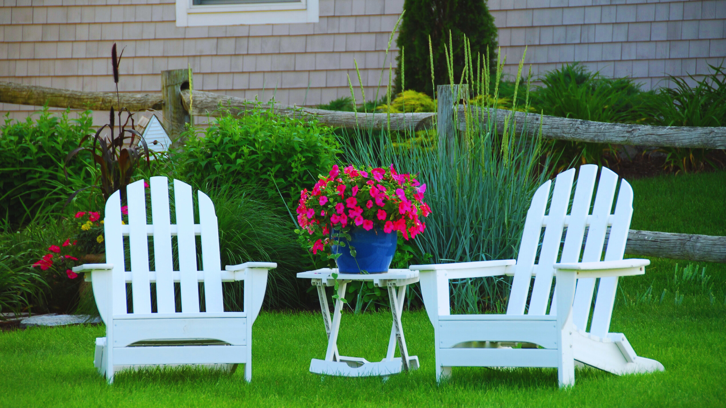 A garden scene with a white table and two white Adirondack chairs on a green lawn, with a blue pot of bright pink flowers on the table. Behind the furniture, there are lush green plants and a wooden fence.
