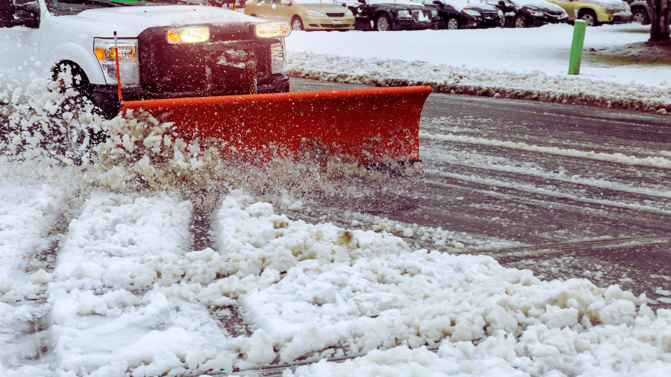 A snowplow truck clearing snow from a parking lot, with snow-covered cars in the background.