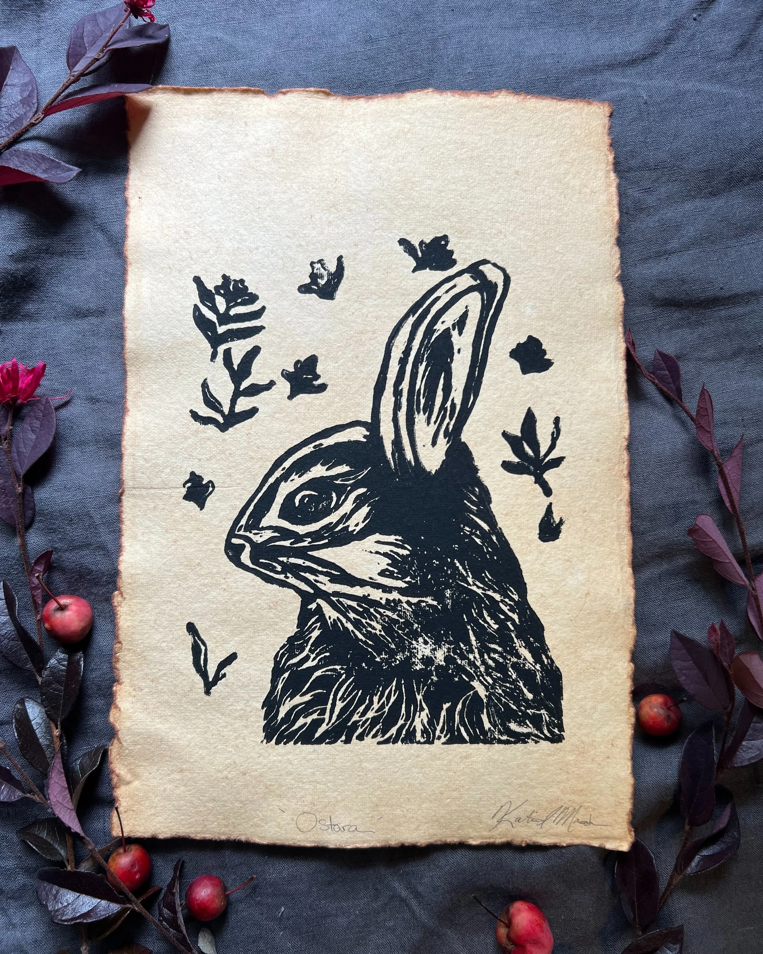 A hand carved, block print of a rabbit with one ear up, surrounded by small leaves and wisps, on handmade paper.