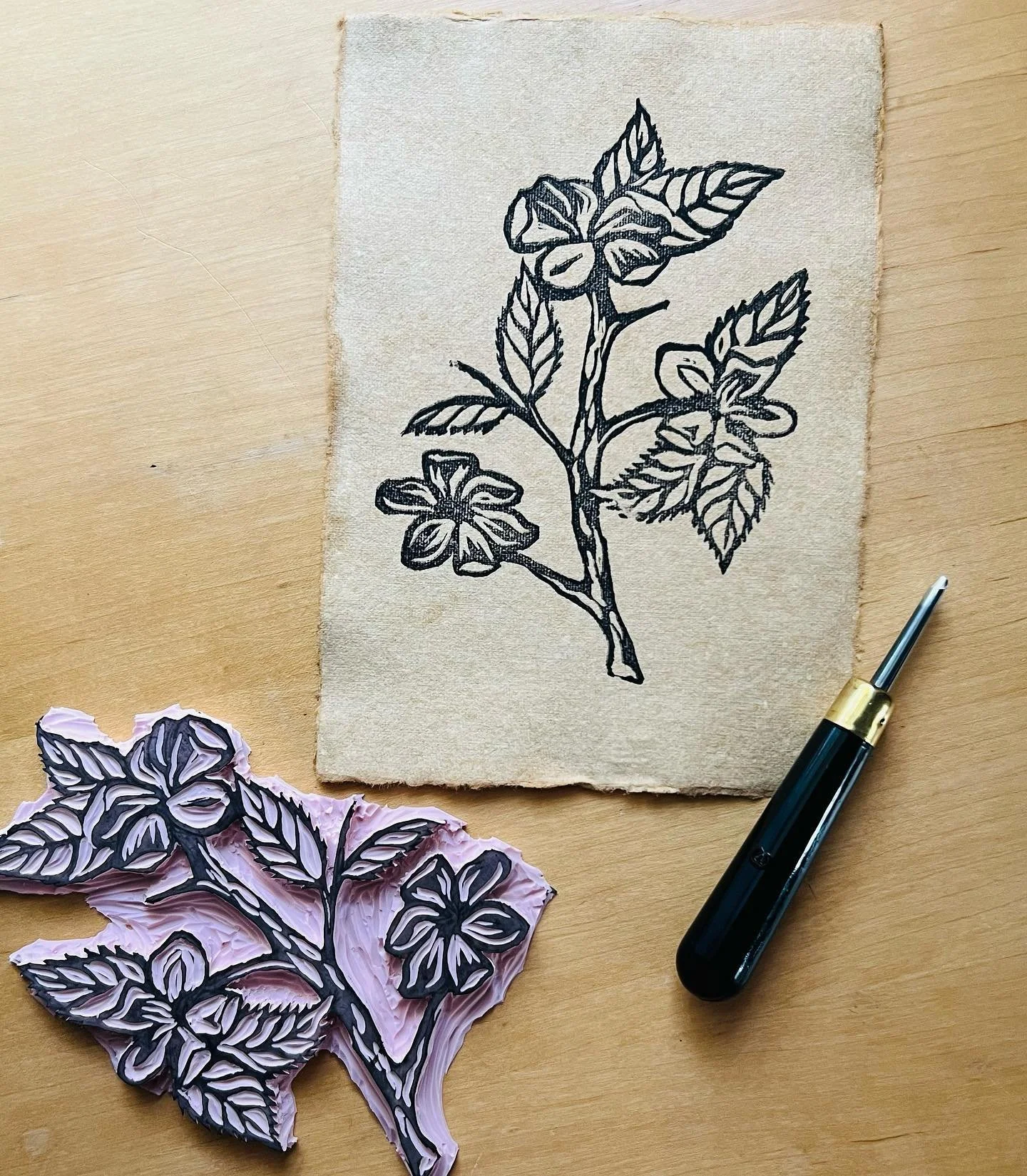 A rubber stamp with an apple blossom carving. A hand stamped print sits next to the stamp of the apple blossom.  A carving tool also sits next to the stamp and print.