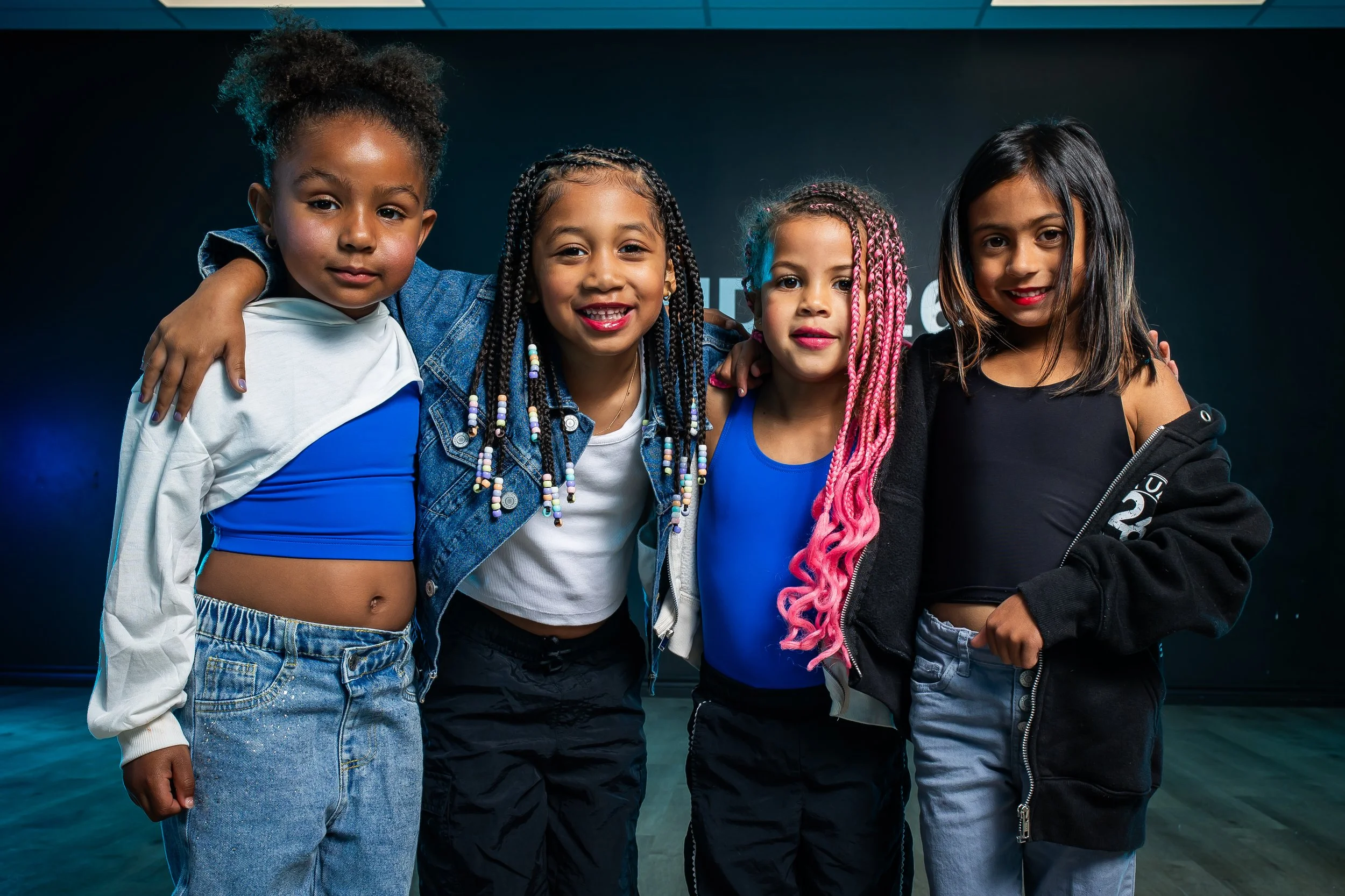 A group of nine ethnically diverse children sitting and standing on a brown sofa, smiling, in a room with a dark wall that has the words 'STUDIO 26' and 'Connection, Community, Culture, Creativity' written on it.