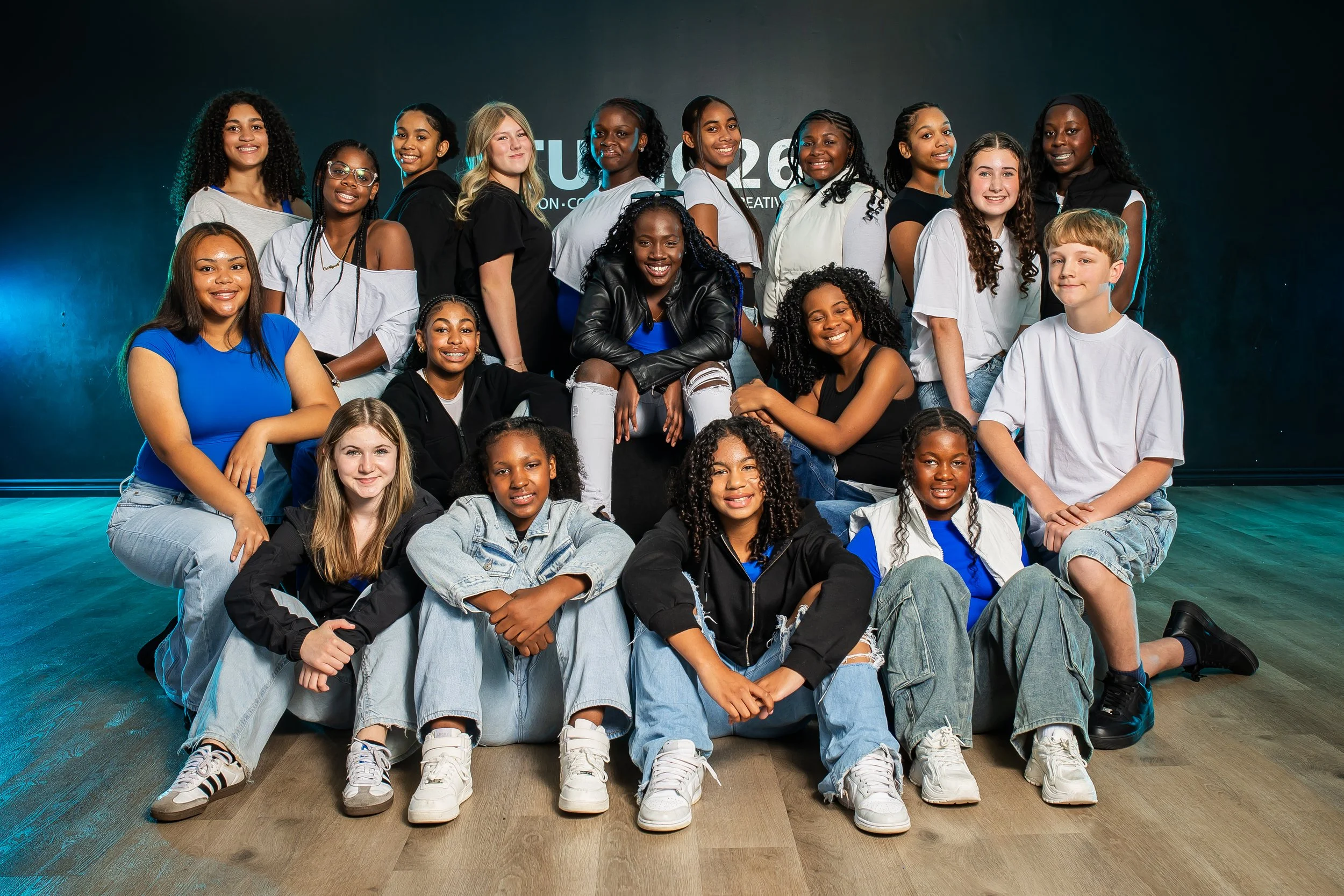 A group of ten diverse young girls poses in a dance studio with dark background and pink and blue lighting. They are dressed in casual outfits, some sitting and others standing, looking serious.