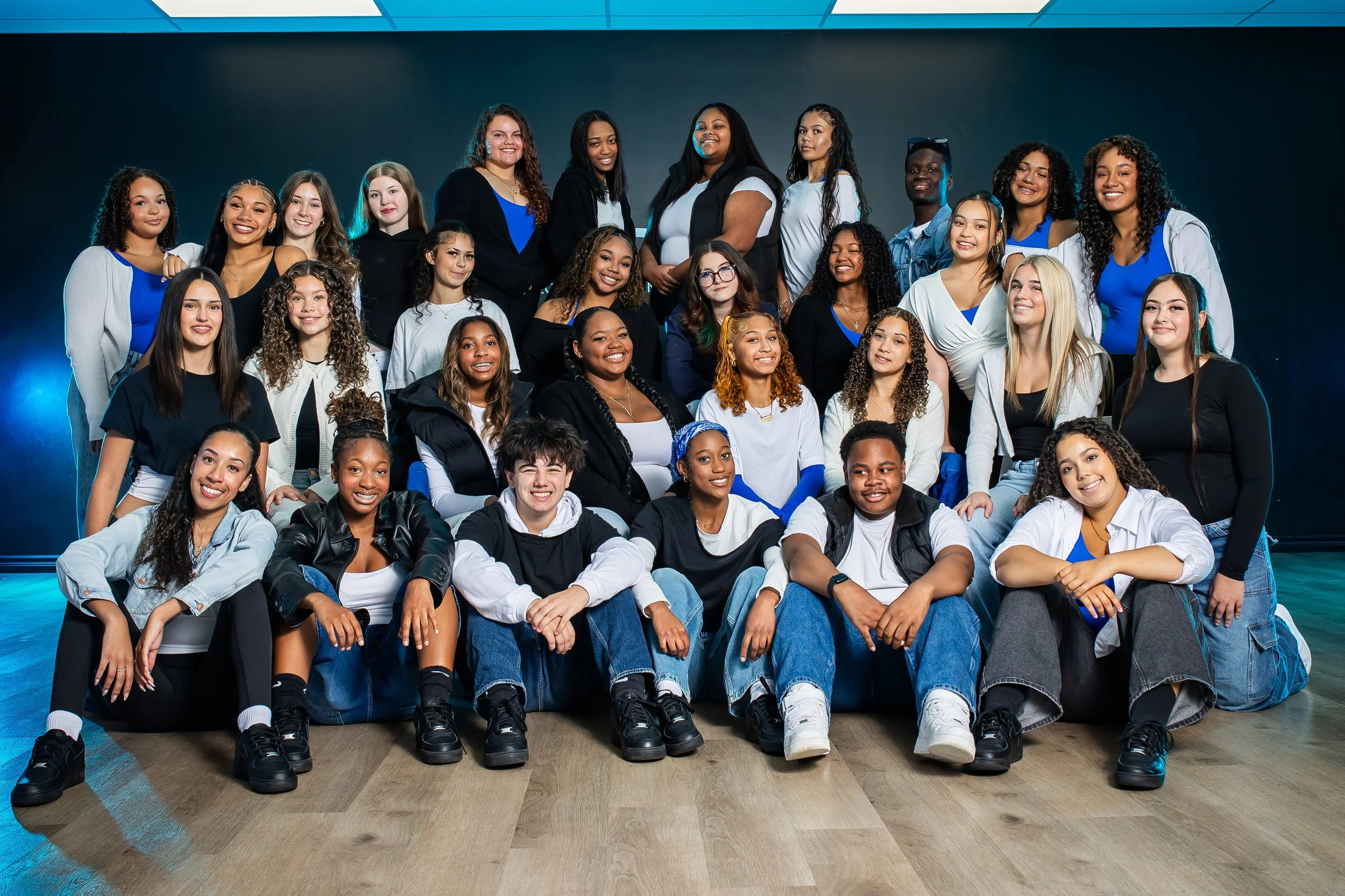 A group of twelve diverse young women standing closely together with their arms around each other's shoulders, smiling at the camera in a dance or fitness studio with a dark wall and purple-lit ceiling.