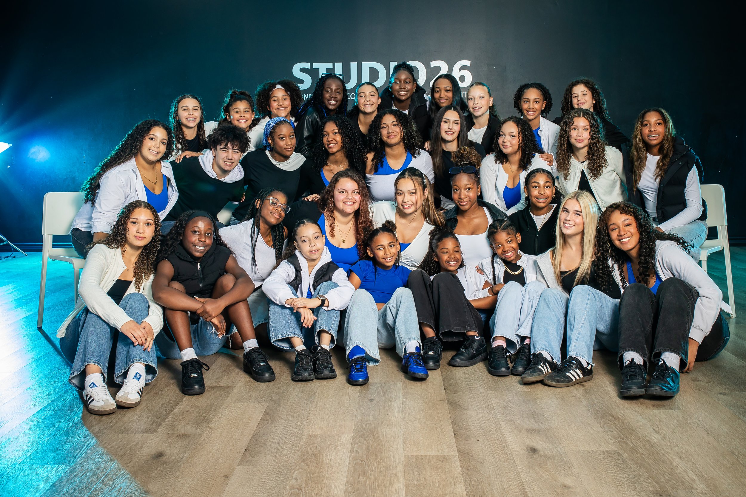 Group photo of diverse women and girls smiling, posing on a stage with a dark background and stage lighting.