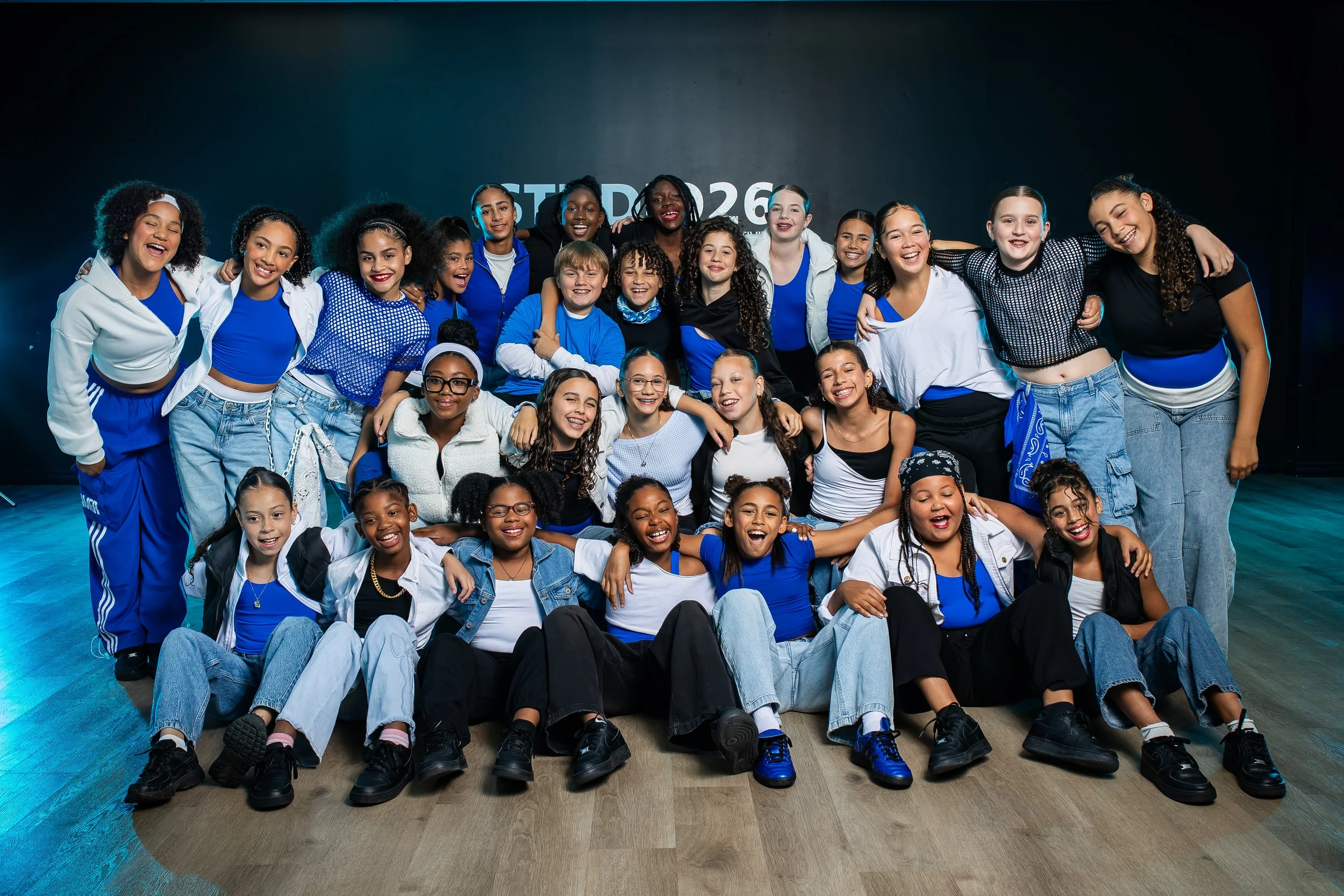 A group of twelve young girls standing together on a dance studio floor, leaning on each other with arms around shoulders, in front of a dark backdrop with 'STUDIO26' written on it. The girls are dressed in casual sportswear and sneakers, with some wearing white vests, puffer jackets, or patterned pants, and have various hairstyles.