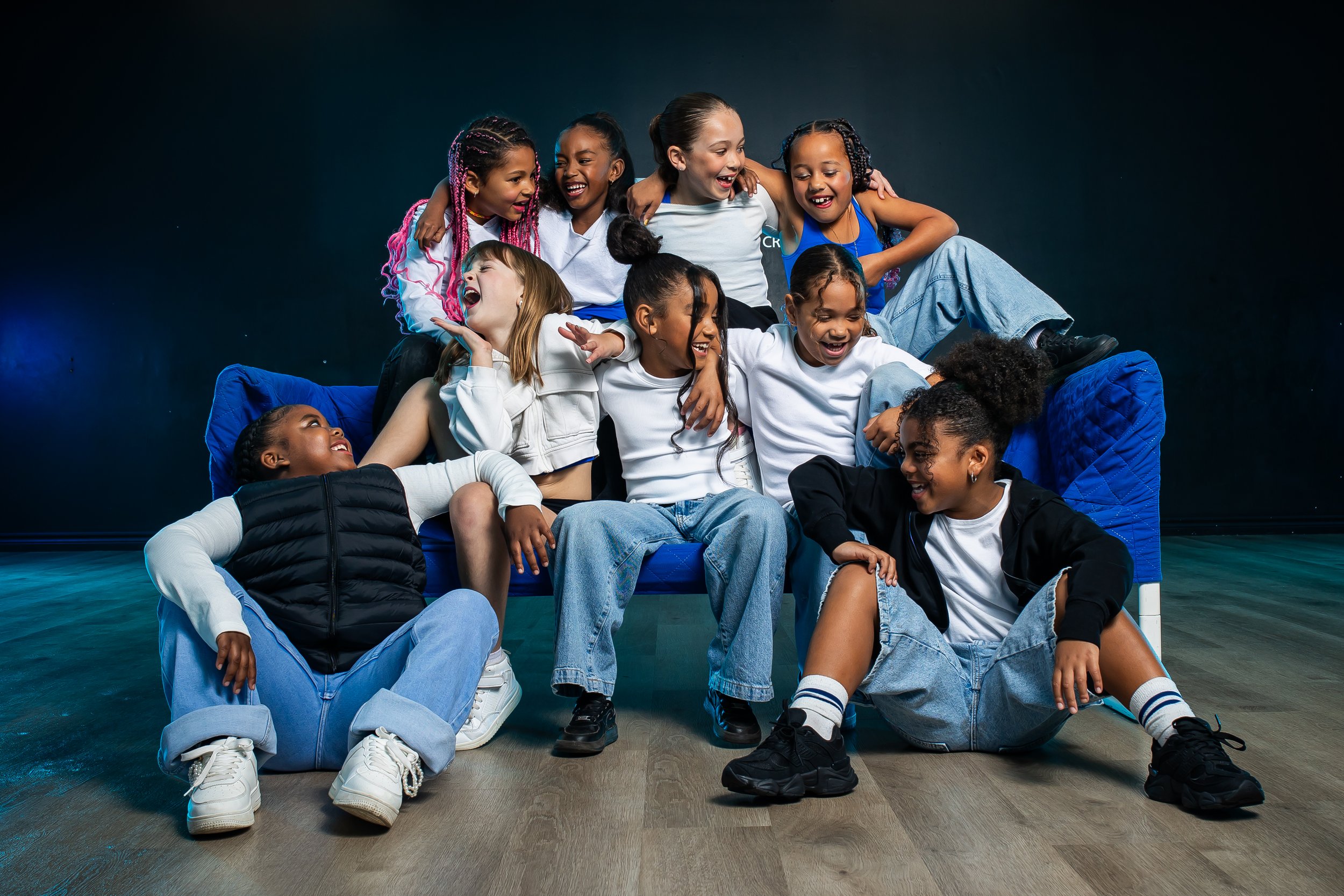 Group of 13 diverse children posing on stage, some standing and some sitting, wearing casual clothing and white sneakers.