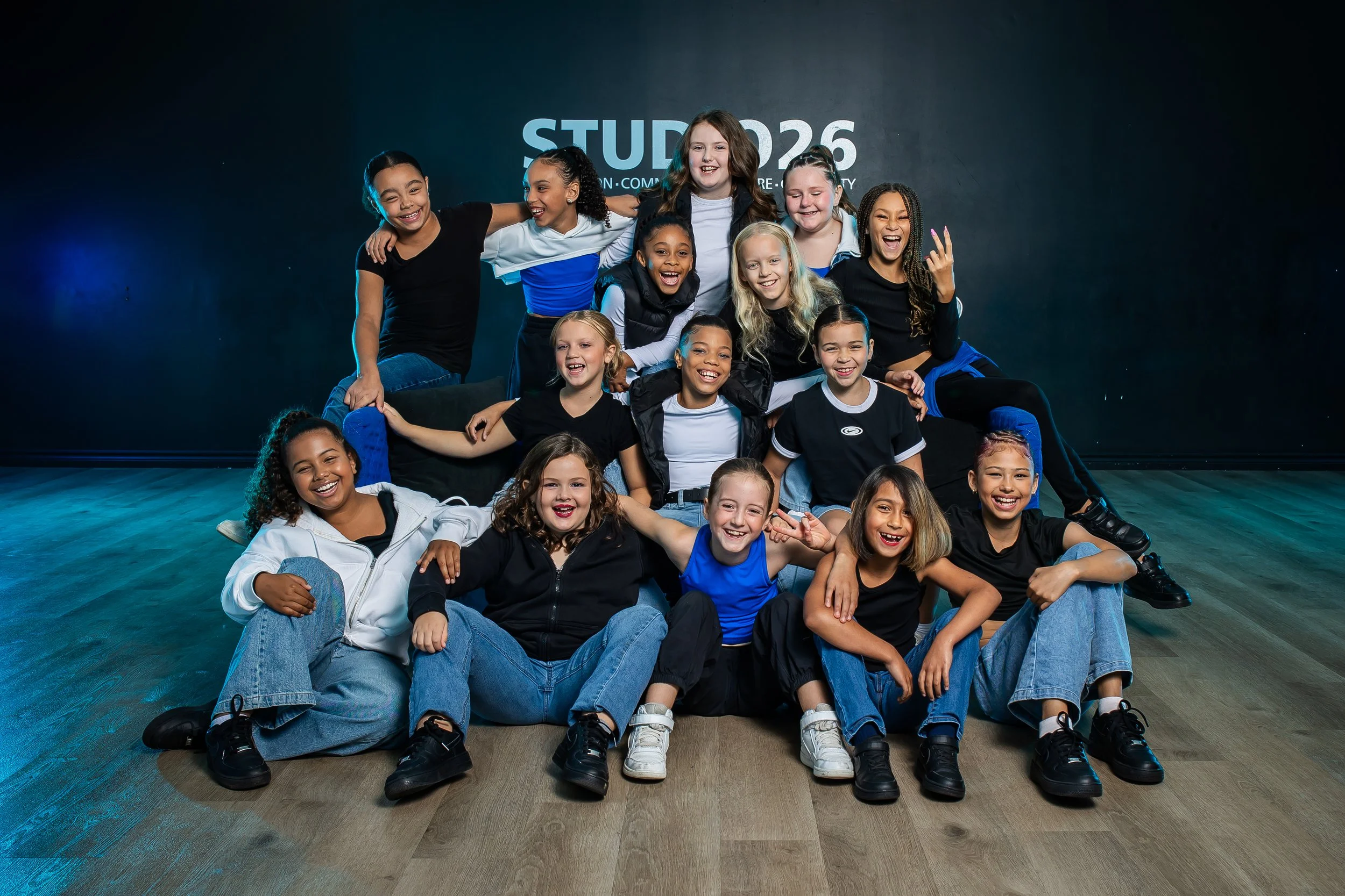 A diverse group of young children posing together on a stage with a dark background and pink lighting.