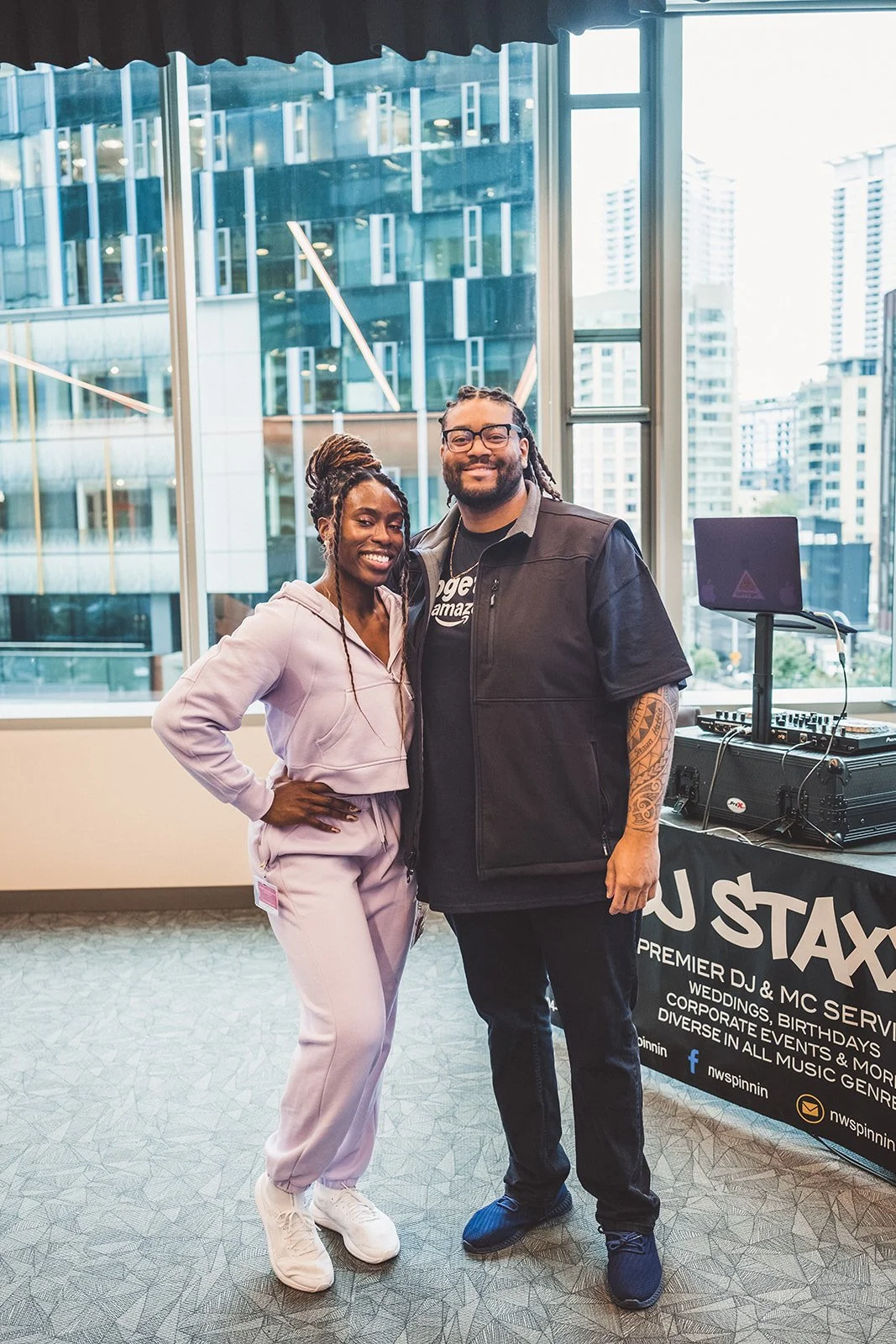 Two smiling people posing in front of a DJ setup at an indoor event with large windows showing city buildings outside.