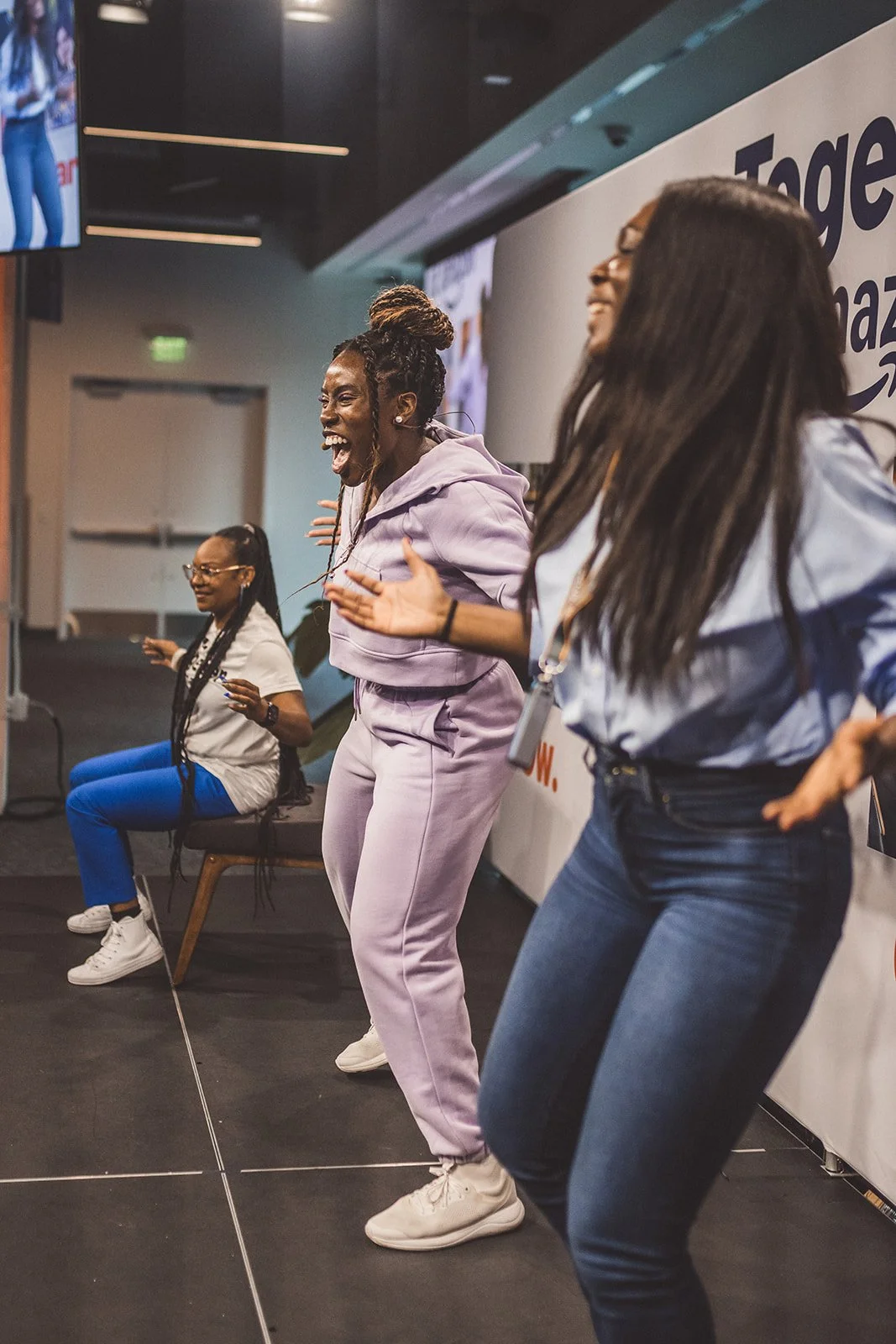 Three women enthusiastically dancing or singing on stage at an Amazon event, with two women sitting in the background.