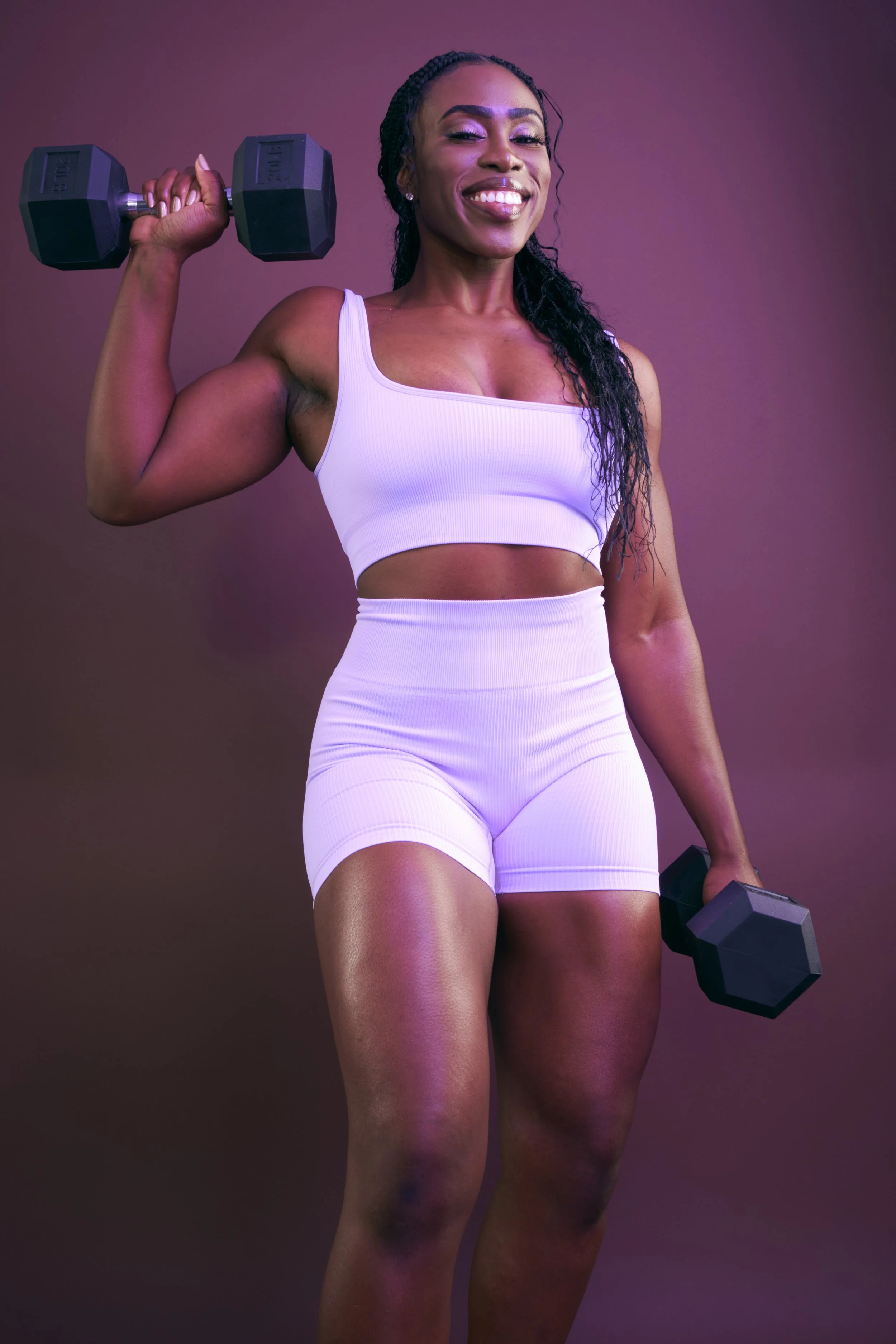 A woman in white athletic wear holding a dumbbell in each hand, smiling, with a purple background.