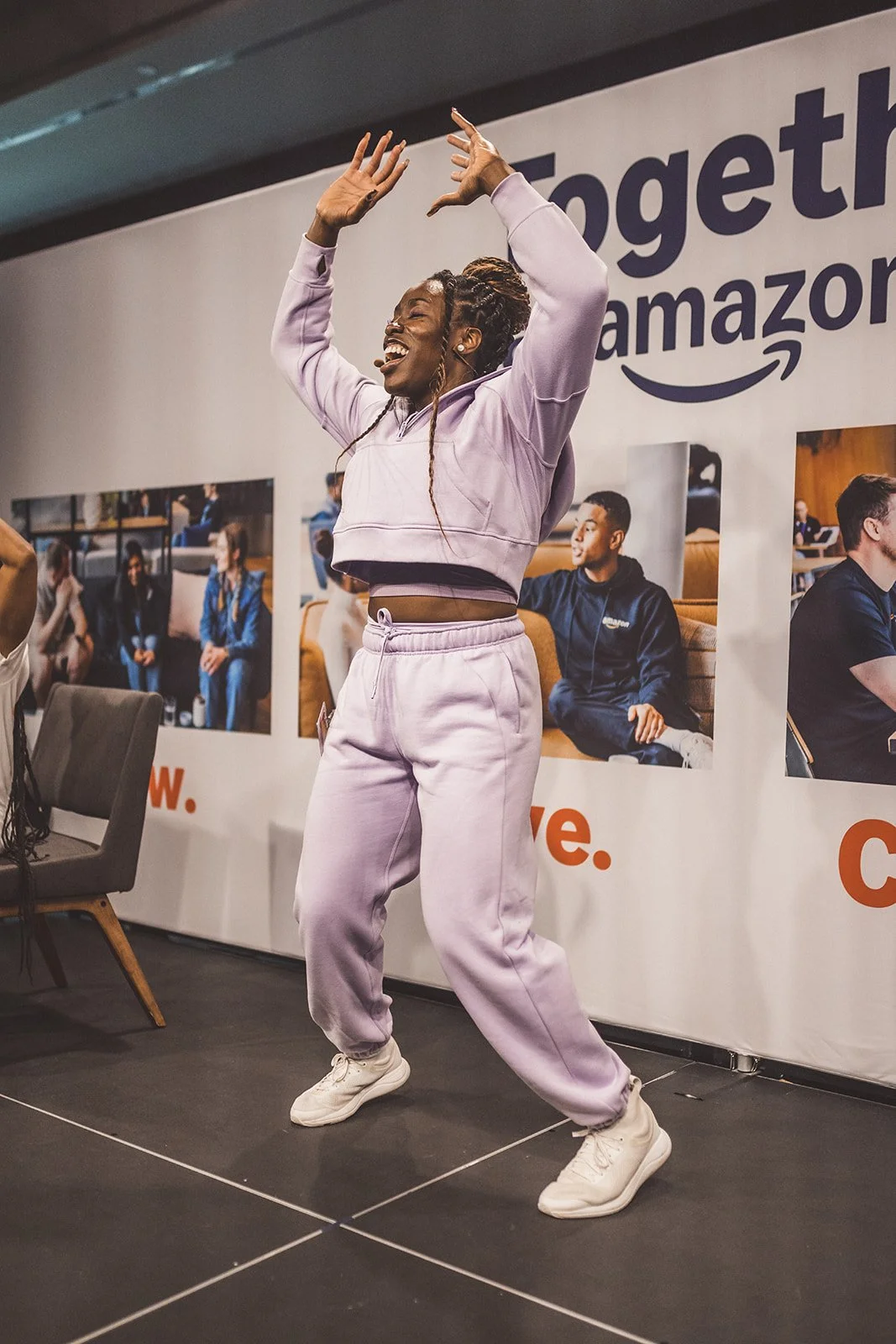 A woman dancing happily on stage at an Amazon event, wearing a light purple tracksuit and white sneakers, with a large Amazon banner in the background.