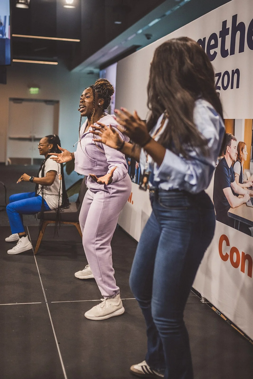 Three women dancing and clapping at an event, with a large banner in the background featuring the Amazon logo.
