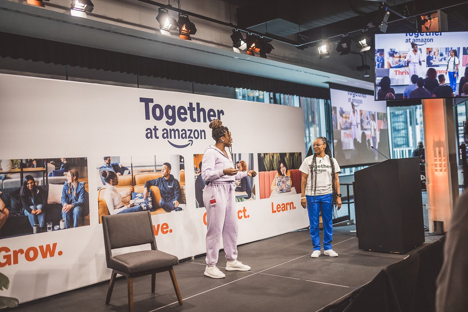 Two women standing on a stage at an Amazon event, with a large banner behind them that reads "Together at amazon" and photos of people working in an office. The women are engaged in conversation, one in a white sweatshirt and sweatpants, the other in