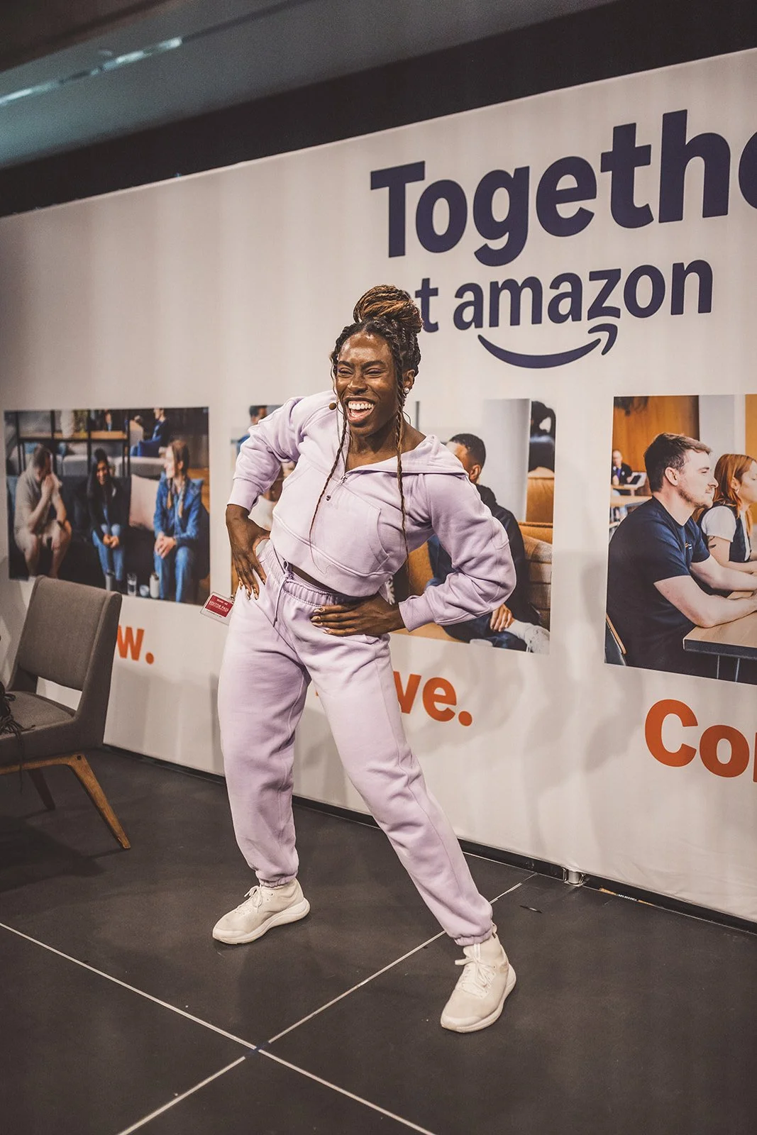 A woman in a lavender matching tracksuit and white sneakers is smiling and posing in front of an Amazon banner during an event.
