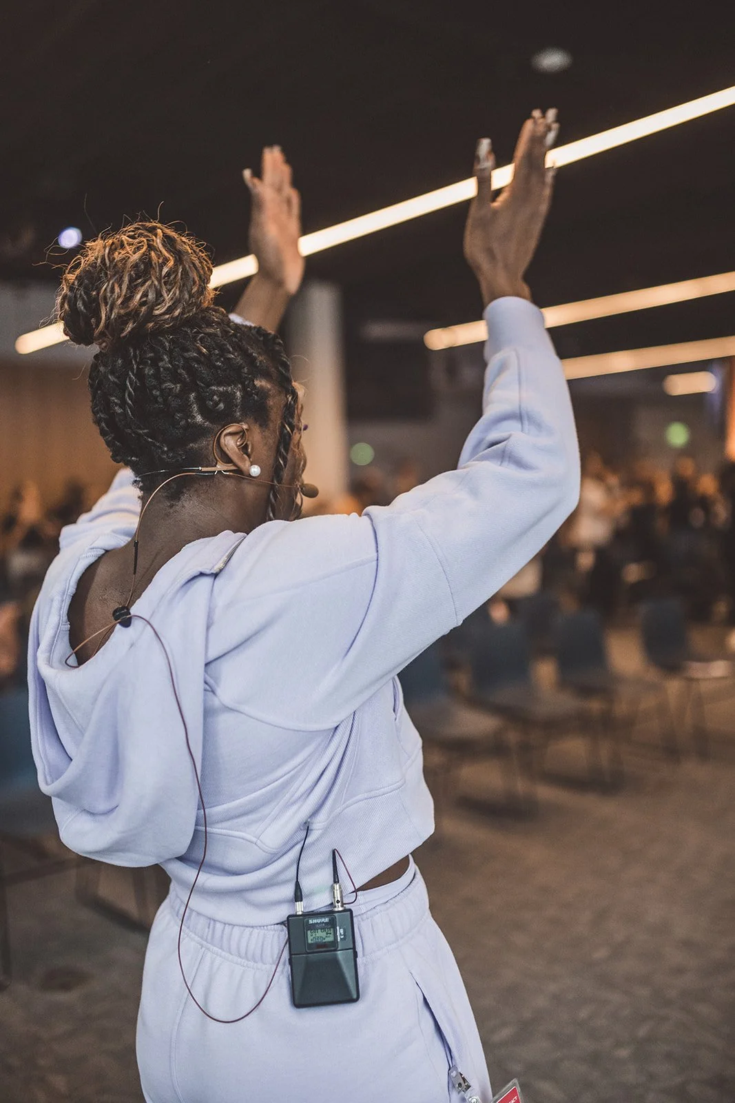 A woman with braided hair and a headset raises her hands in the air at an indoor event with a blurred audience in the background.