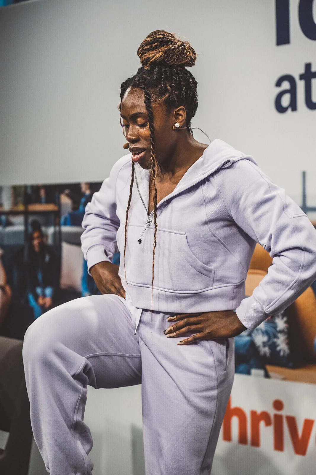 A woman with styled dreadlocks in a bun, wearing a light purple hoodie and matching sweatpants, speaking into a headset microphone and demonstrating a move with her hand on her hip.