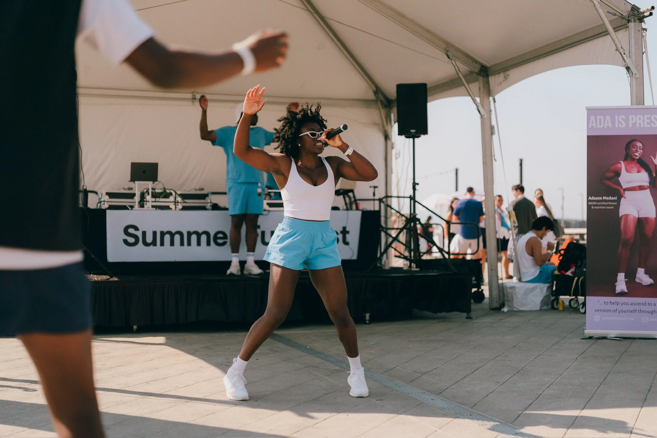 A woman in white tank top, light blue shorts, and white sneakers dancing and singing into a microphone under a large outdoor tent, with a DJ on a stage and people in the background.