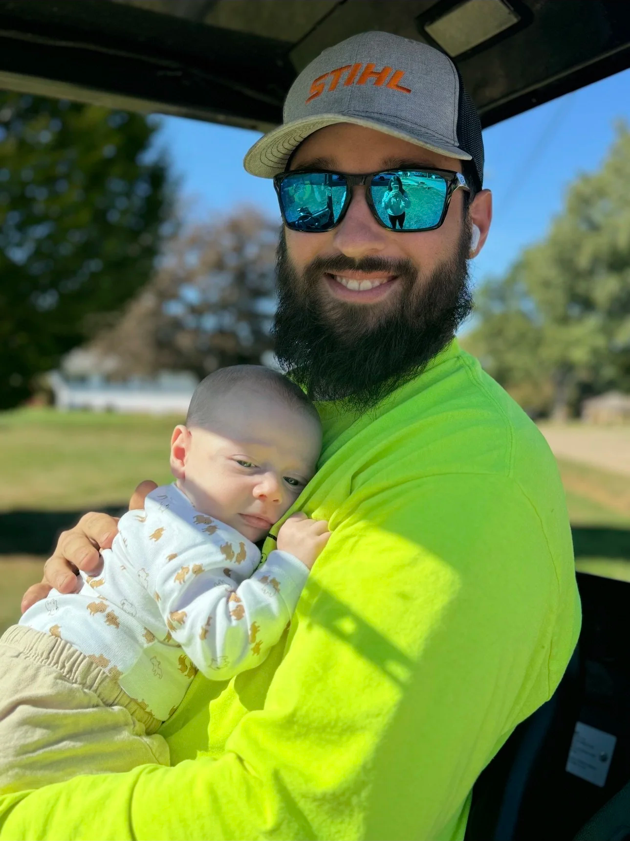 A bearded man wearing sunglasses, a gray cap with orange logo, and a bright yellow-green shirt, smiling while holding a young baby with short hair, dressed in a white onesie with animal prints, on his shoulder outdoors on a sunny day.