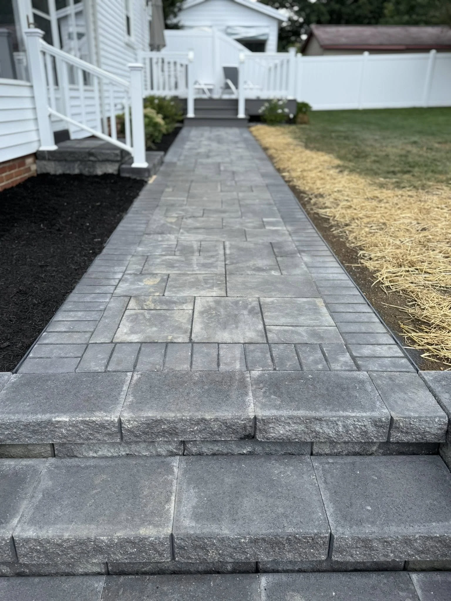 A gray stone walkway with steps leading up to a small porch, surrounded by black mulch and a grassy yard with straw-covered soil, white fencing, and outdoor furniture.