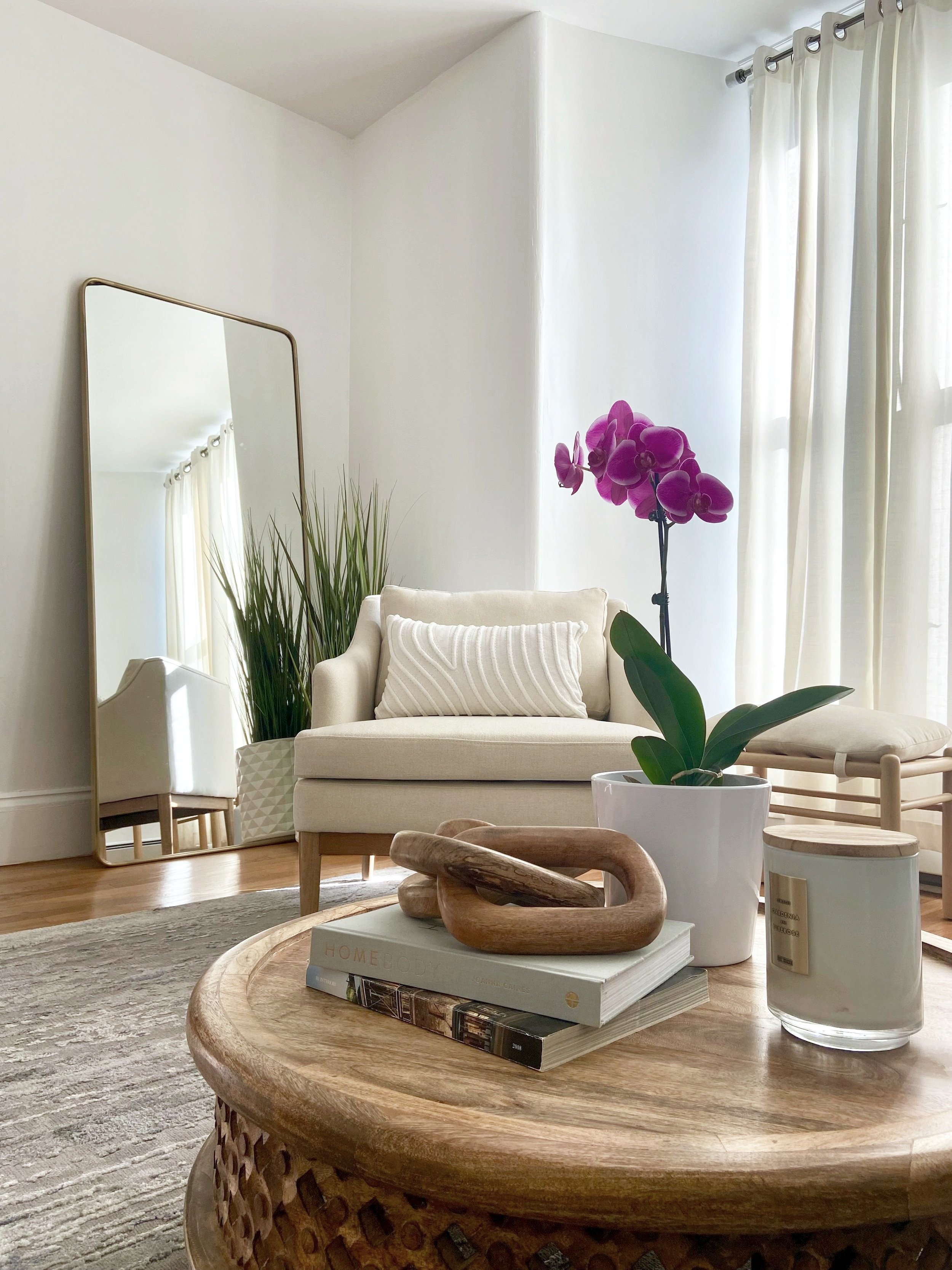 Living room with a beige sofa, a large mirror, a potted purple orchid, a tall green plant, a wooden coffee table with books and a wooden sculpture, and a window with white curtains.