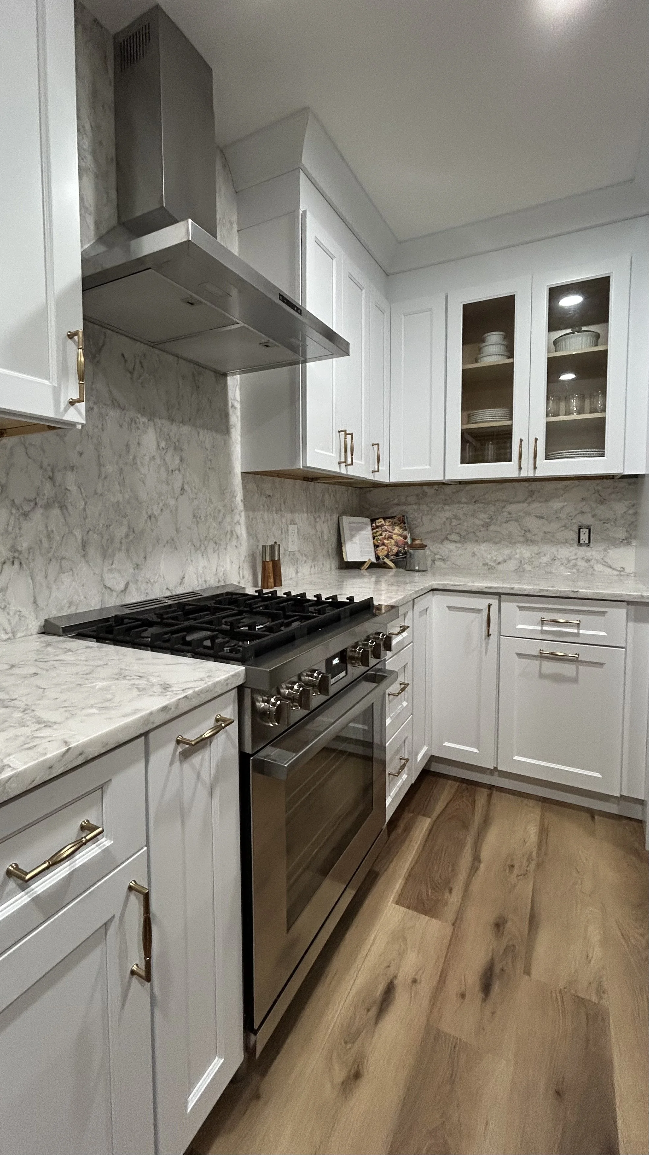 A modern kitchen with white cabinets, a marble backsplash, a stainless steel stove, and wood flooring.