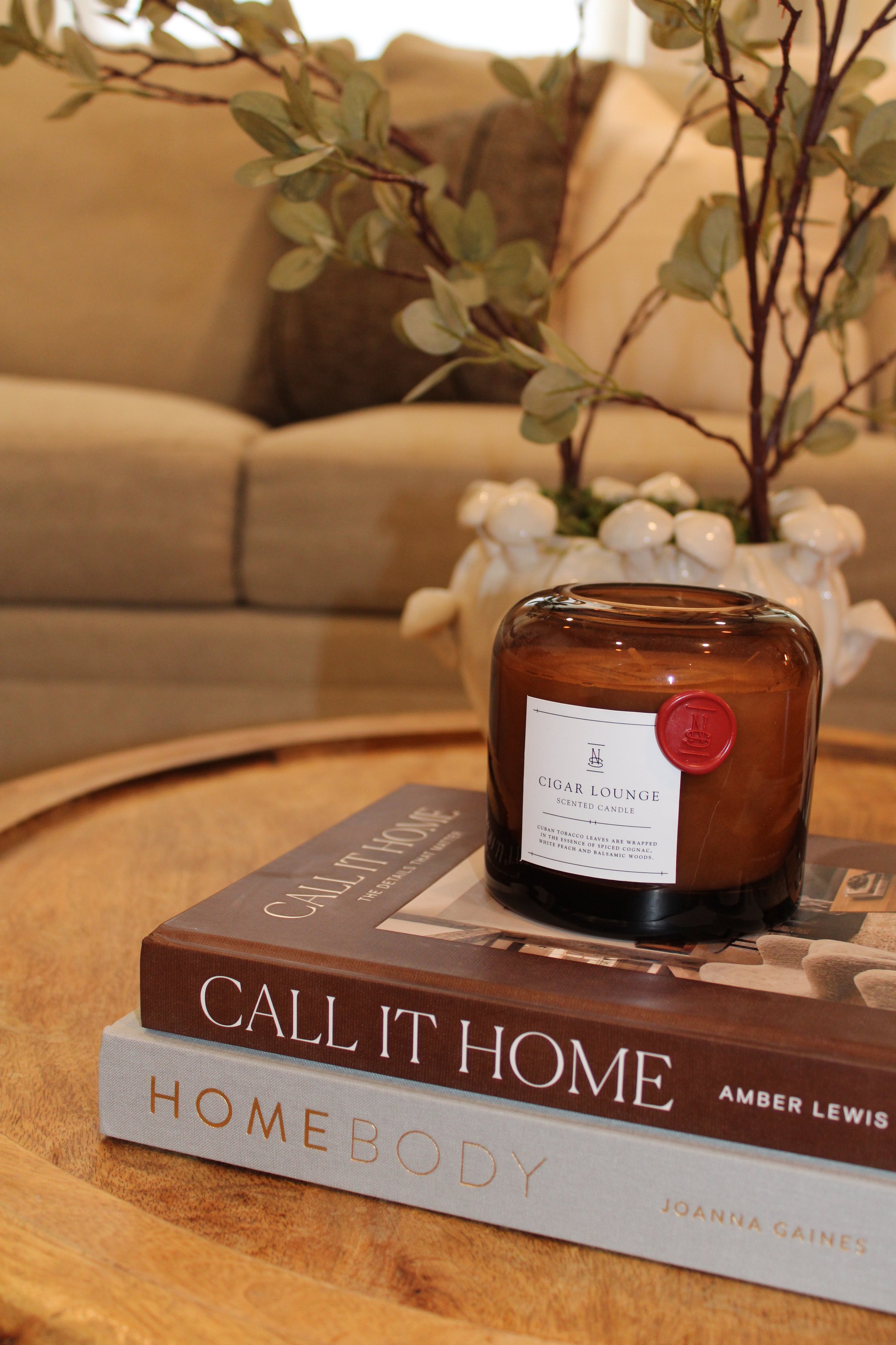 A brown candle labeled 'Cigar Lounge Scented Candle' sitting on two stacked home decor books titled 'Call It Home' and 'Homebody' on a wooden table, with a beige sofa and a white vase with green leafy branches in the background.