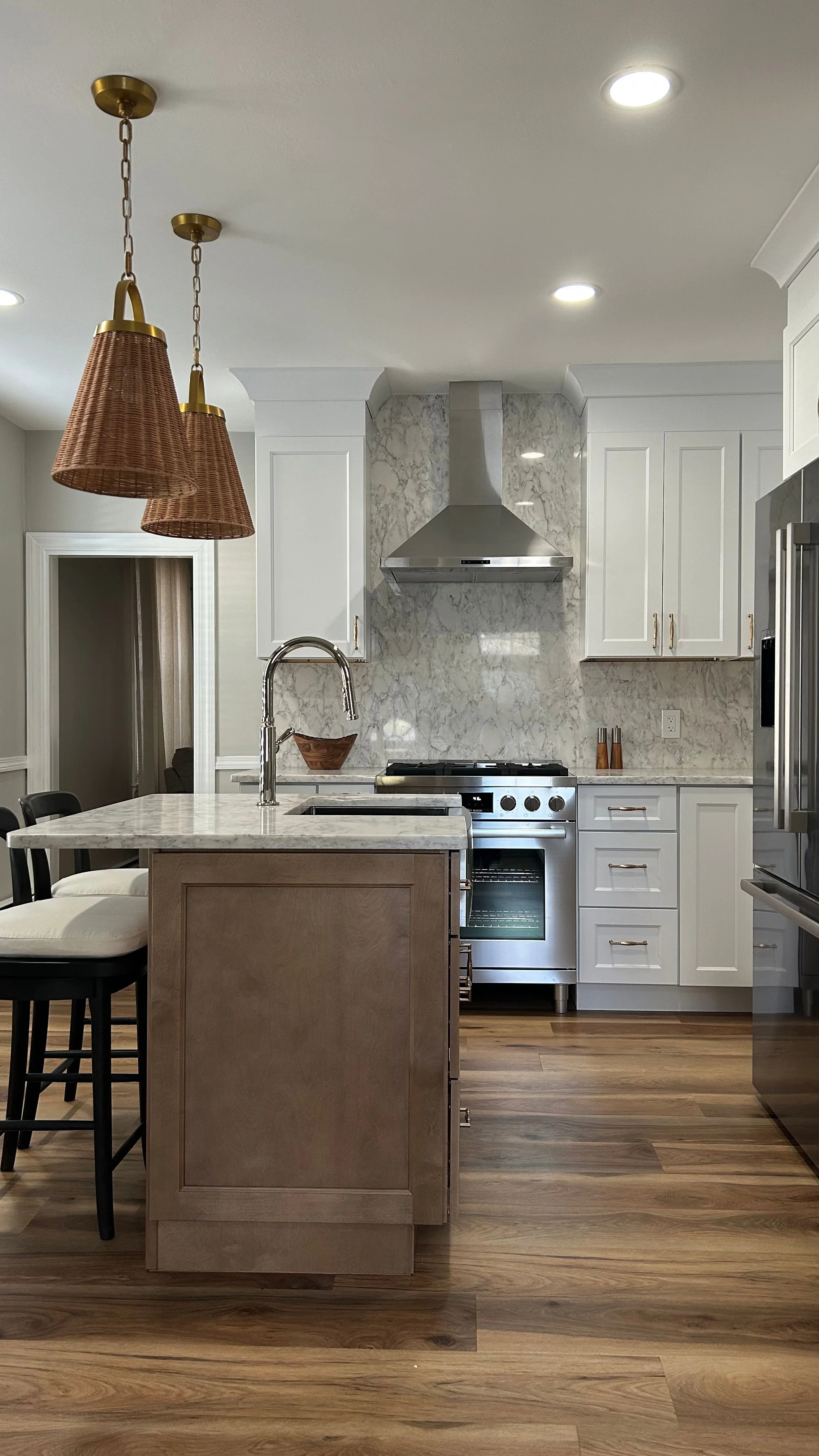 Modern kitchen with white cabinets, stainless steel stove and refrigerator, island with a gray wooden base, marble countertops, and wooden floors. Three wicker pendant lights hang above the island.