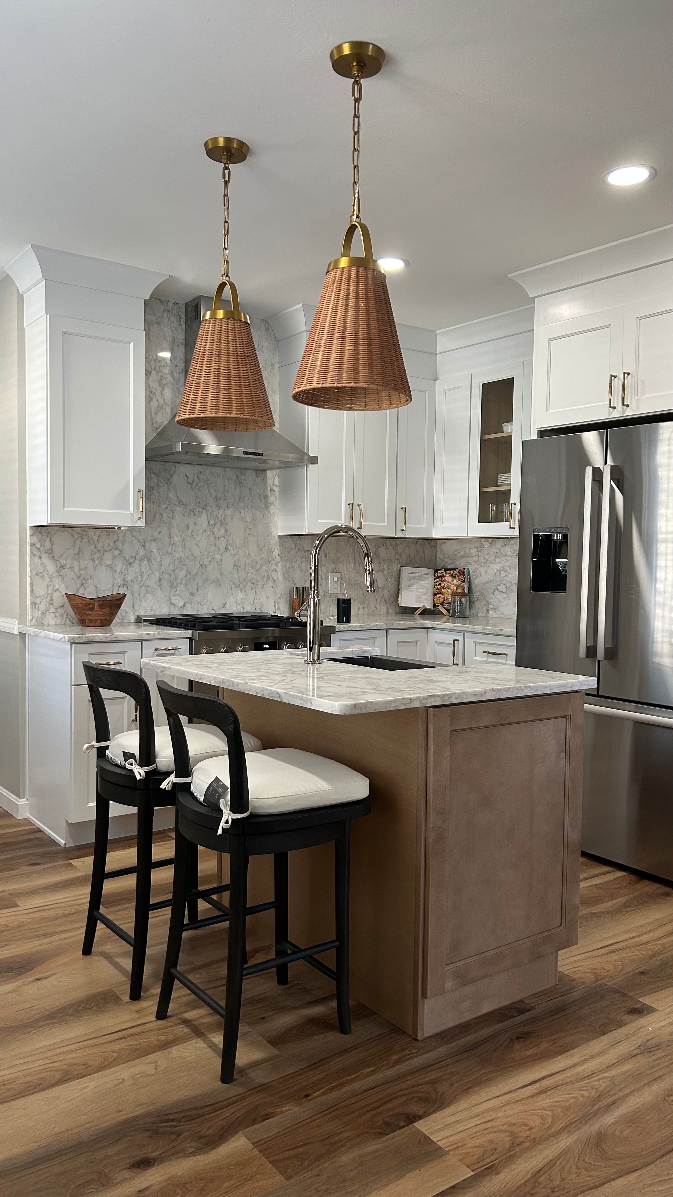 Modern kitchen with white cabinetry, a marble backsplash, a wooden kitchen island with a marble countertop, black chairs with white cushions, stainless steel appliances, and two wicker pendant lights hanging from the ceiling.