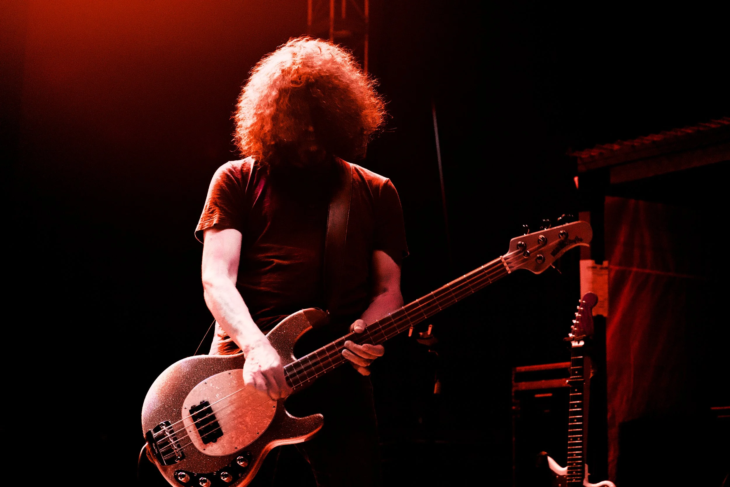 A person with curly hair playing a bass guitar on stage, illuminated by red lighting.