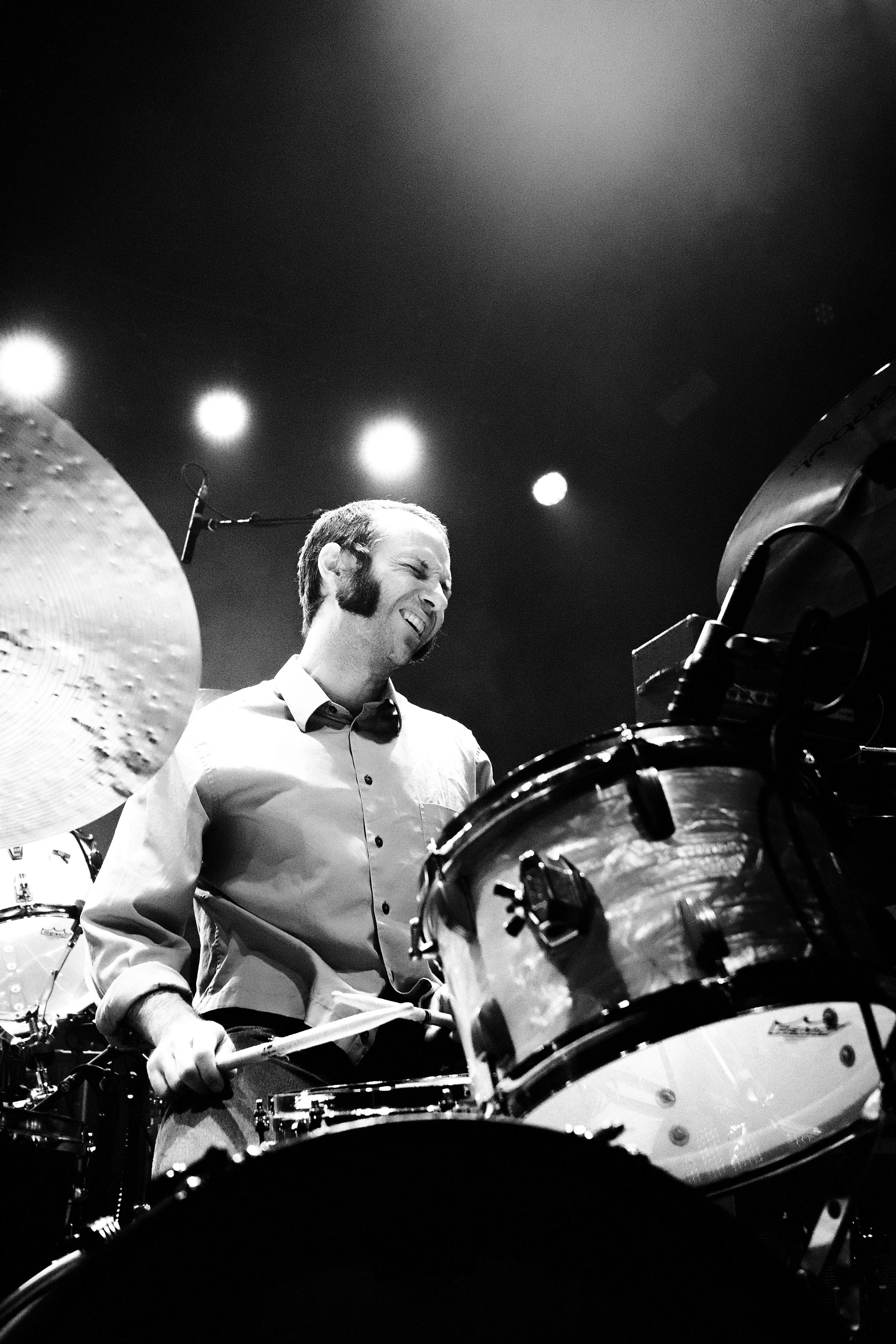 A man playing drums on stage, smiling with eyes closed, with bright lights overhead in black and white.