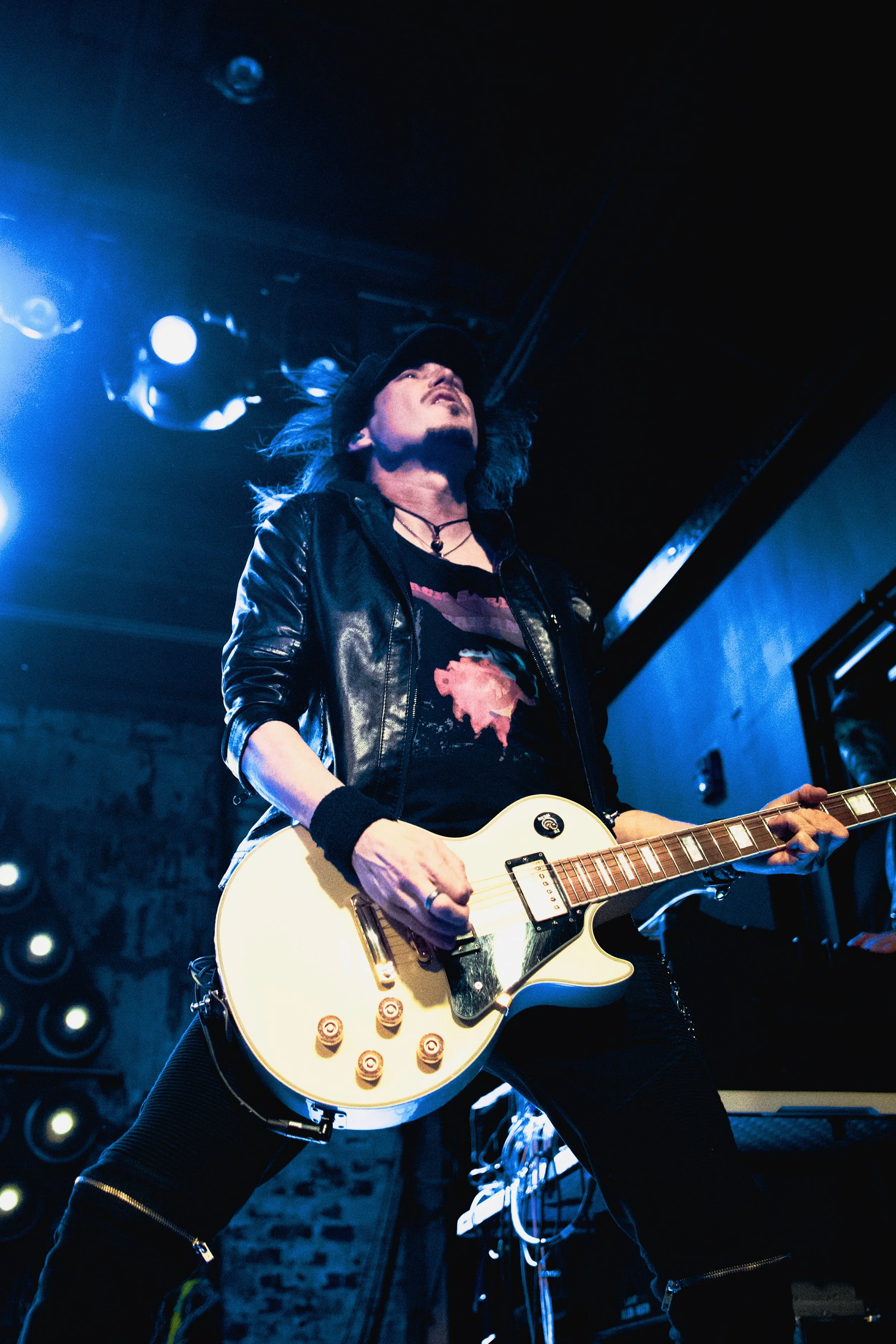 A male guitarist performing on stage under blue lighting, wearing a black leather jacket, a black hat, and playing a white electric guitar.