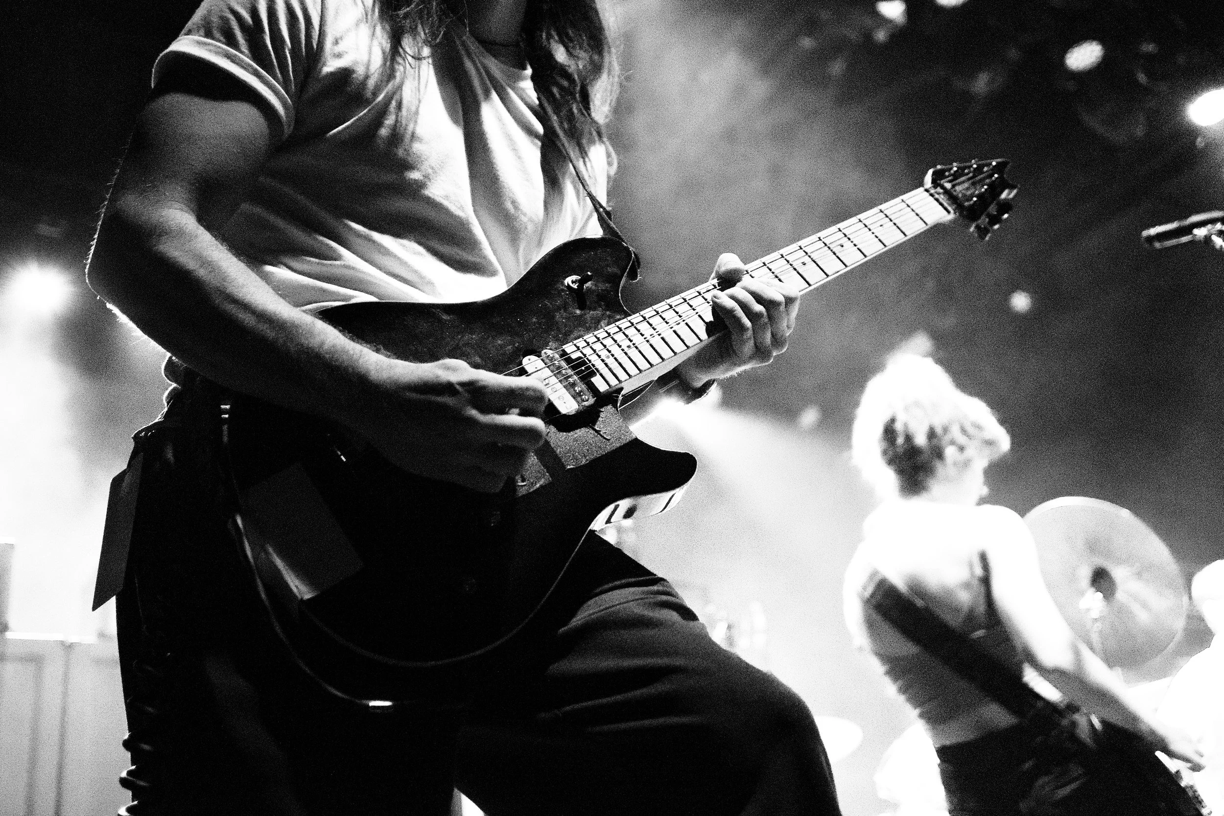 A black and white photo of a musician playing an electric guitar on stage, with another musician playing drums in the background.
