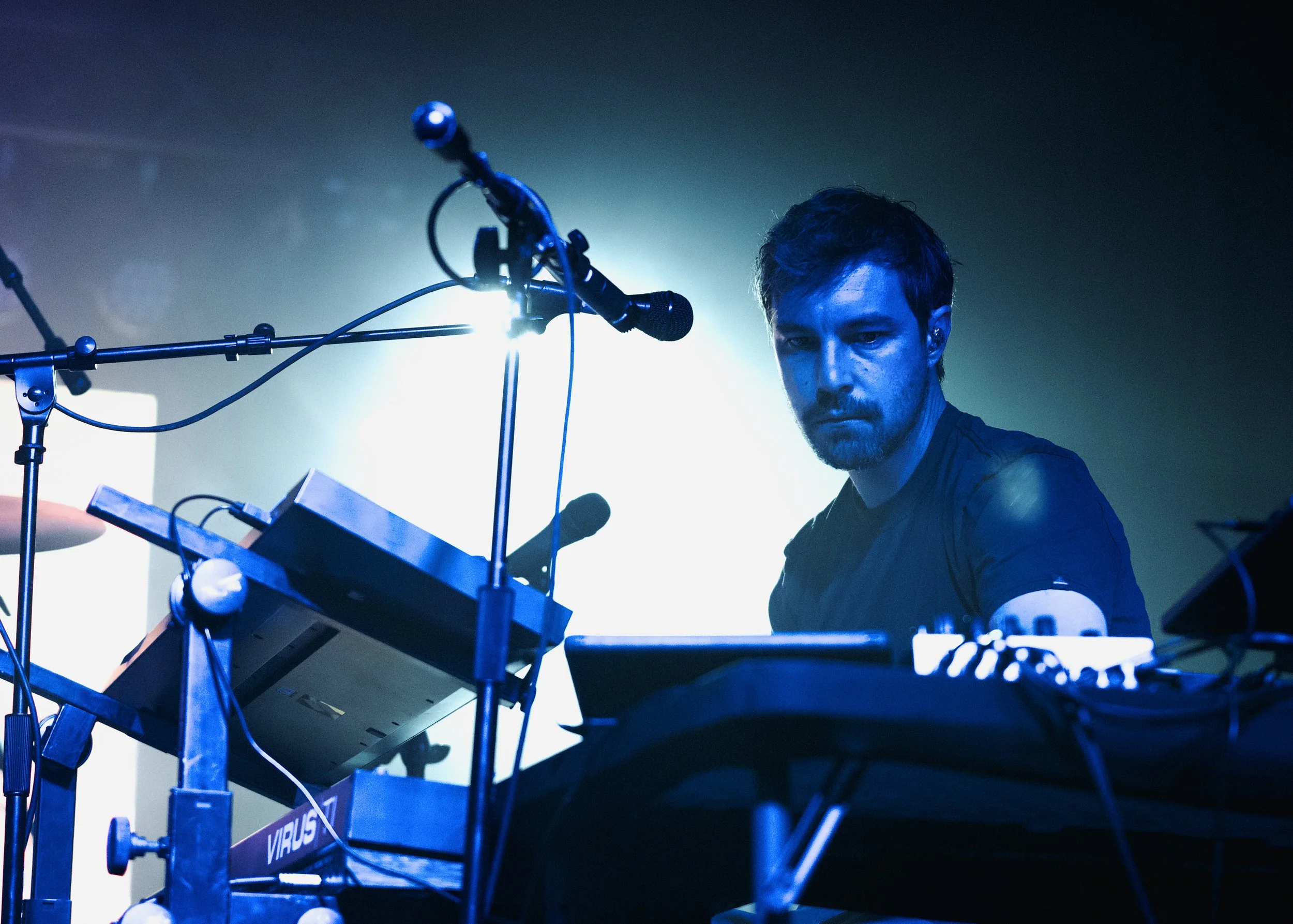 A male musician with a serious expression performing on stage with electronic music equipment, blue and green lighting, and a microphone setup.