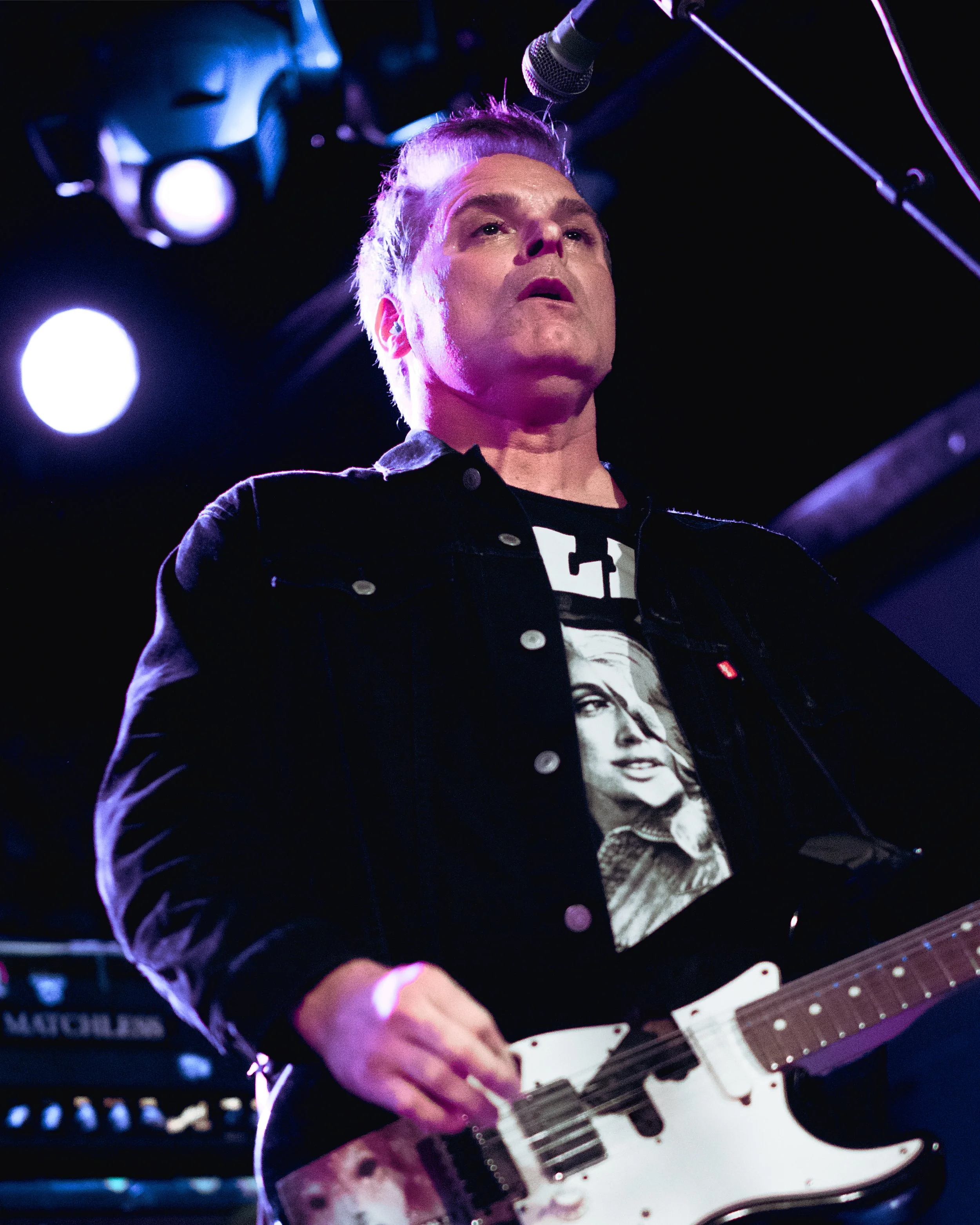 A male musician playing an electric guitar on stage, illuminated by purple stage lights.