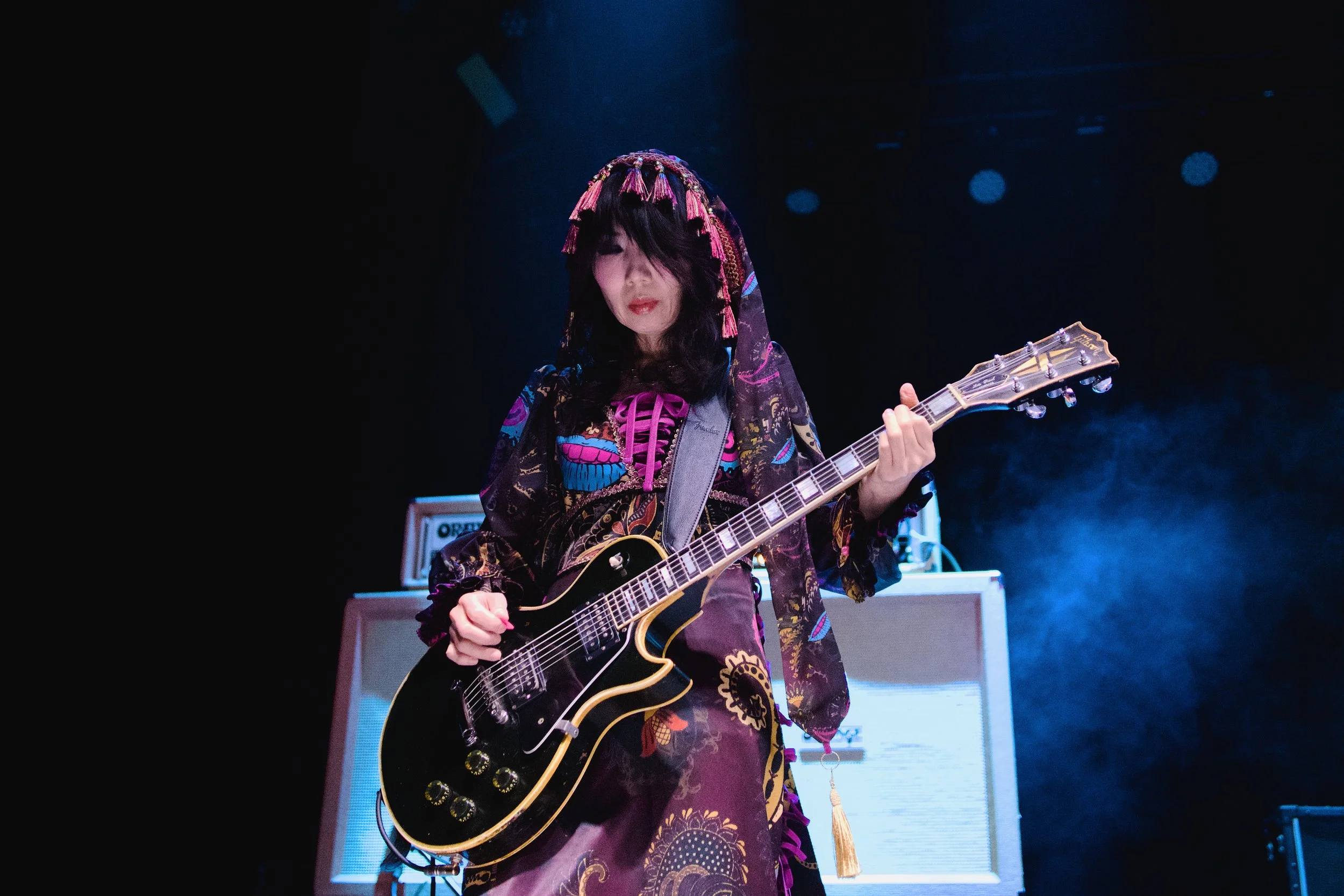 Musician playing an electric guitar on stage with amplifiers in the background, wearing a colorful, embroidered dress and headscarf.