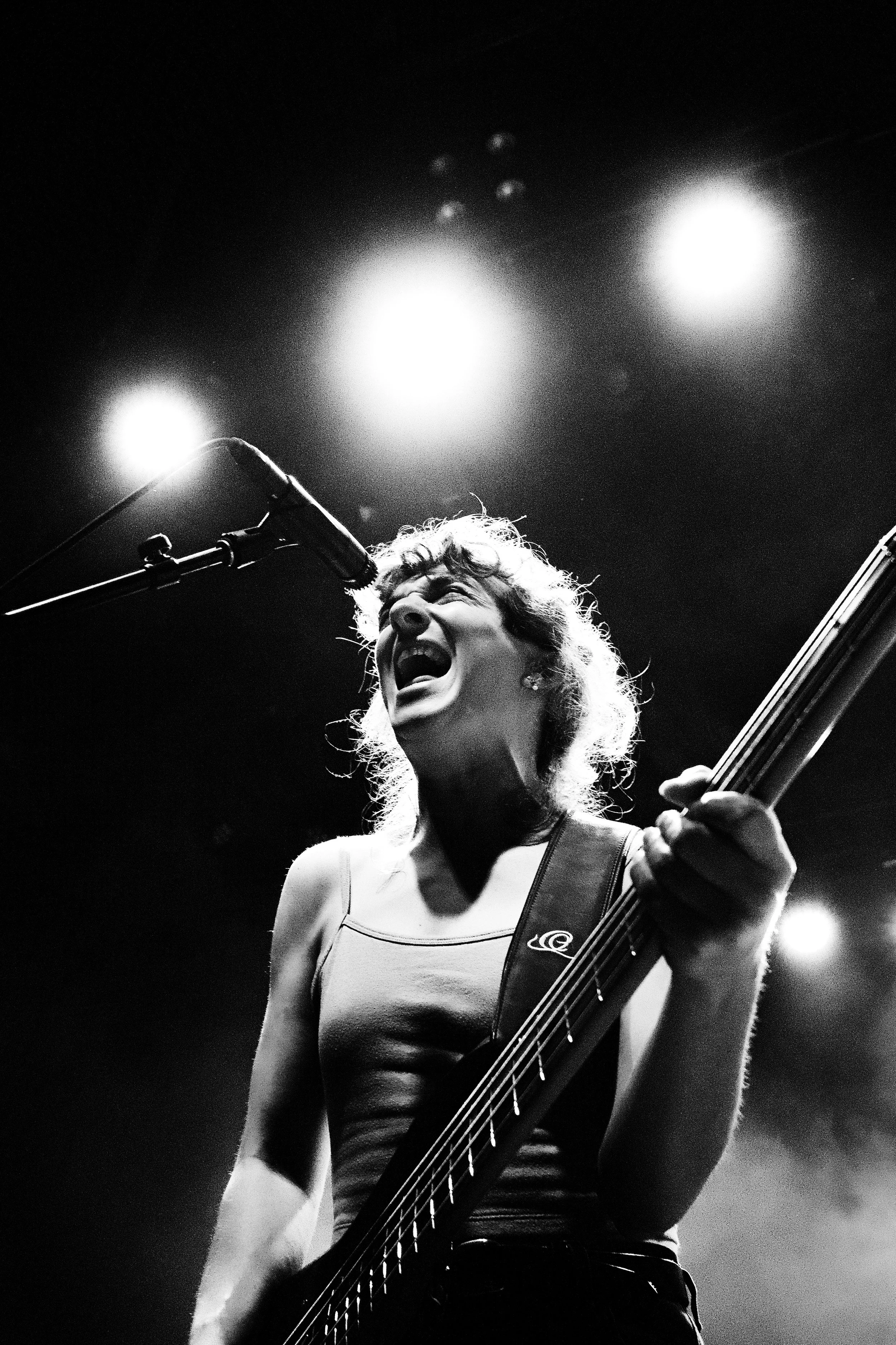 Black and white photo of a woman singing passionately into a microphone while playing an electric guitar on stage, with bright stage lights overhead.