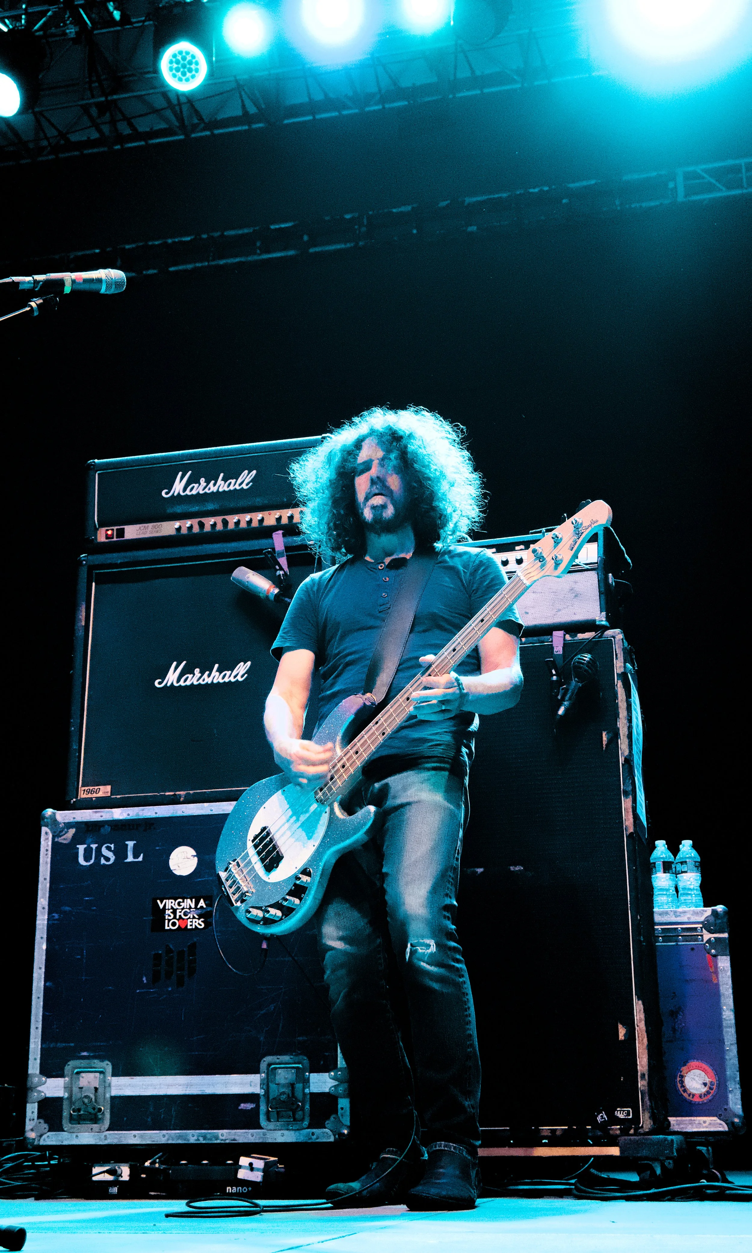 A musician with curly hair playing an electric bass guitar on stage with Marshall amplifiers and stage lights overhead.