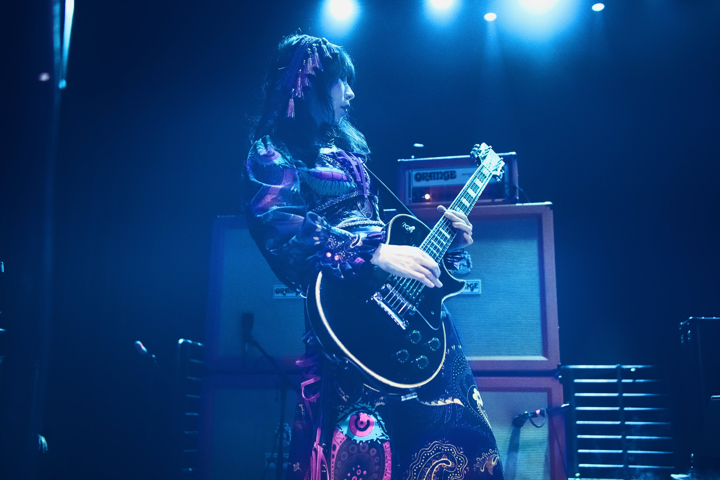 A woman with dark hair and colorful braided extensions playing an electric guitar on stage, illuminated by blue stage lights, with guitar amplifiers in the background.