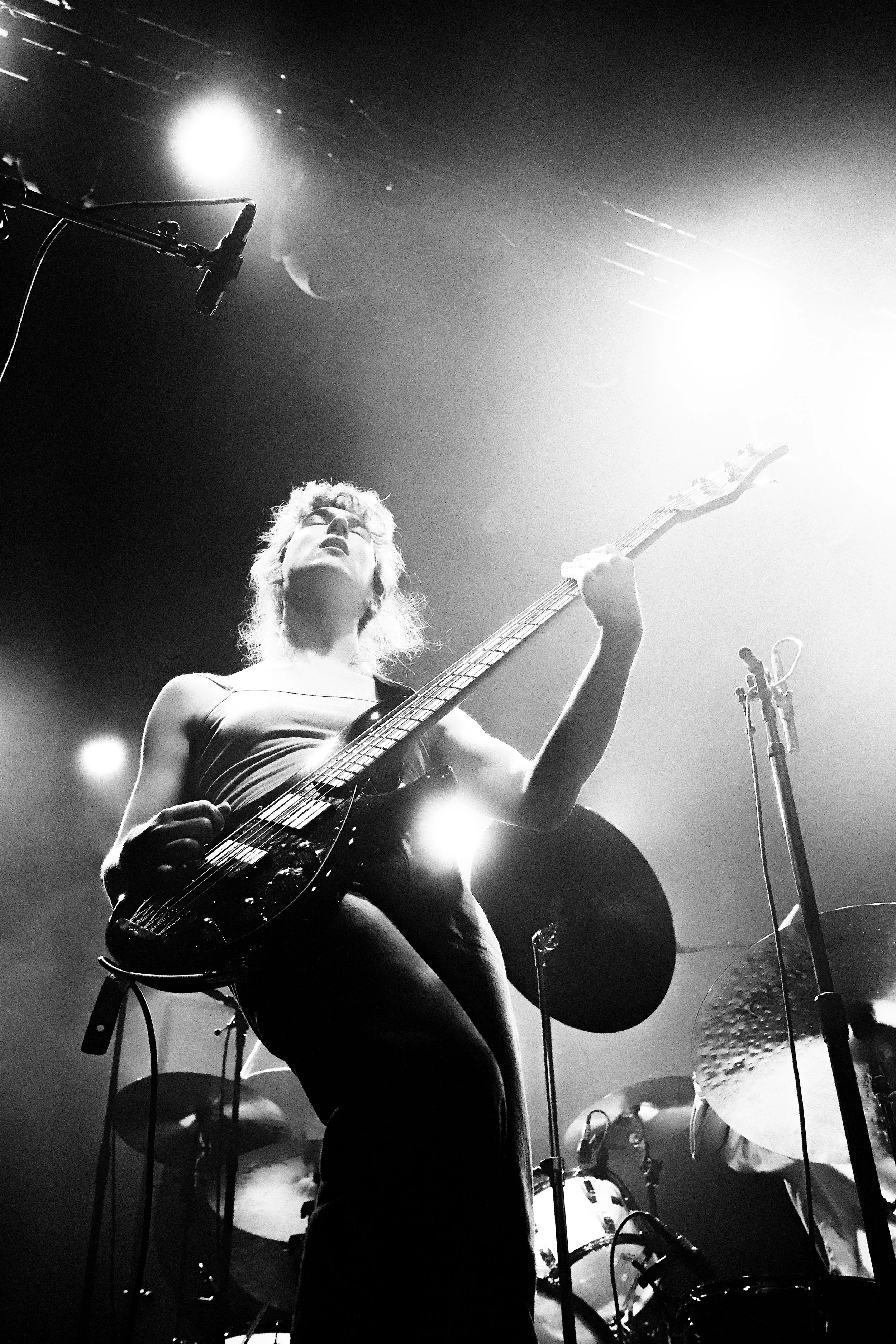 A musician playing an electric guitar on stage with bright stage lights overhead. The scene is in black and white, capturing a dynamic performance.