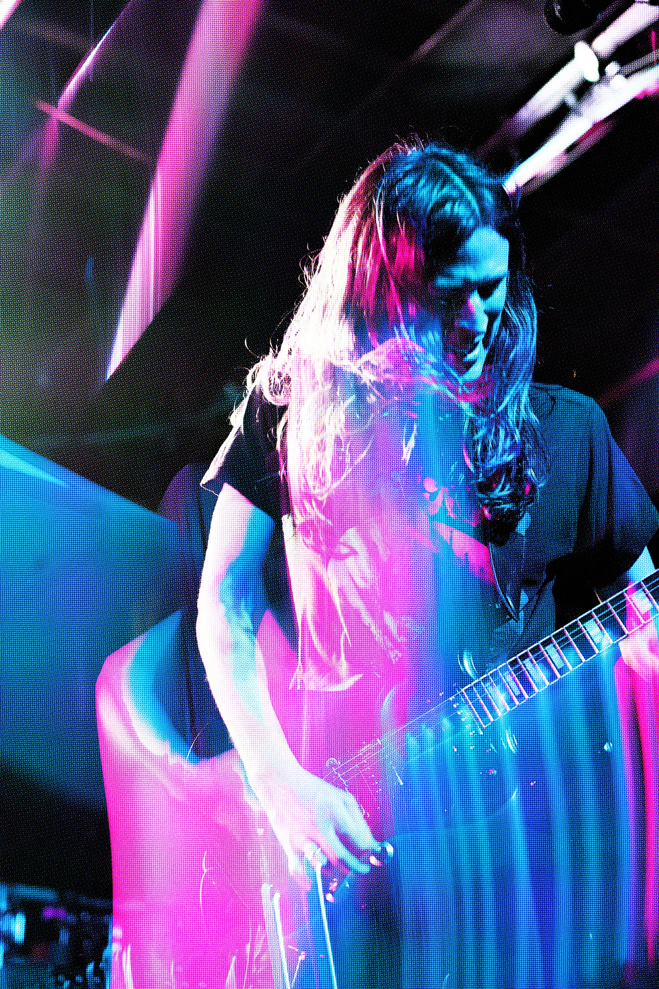 A person with long hair playing an electric guitar in a dark setting illuminated with vibrant pink and blue stage lighting.