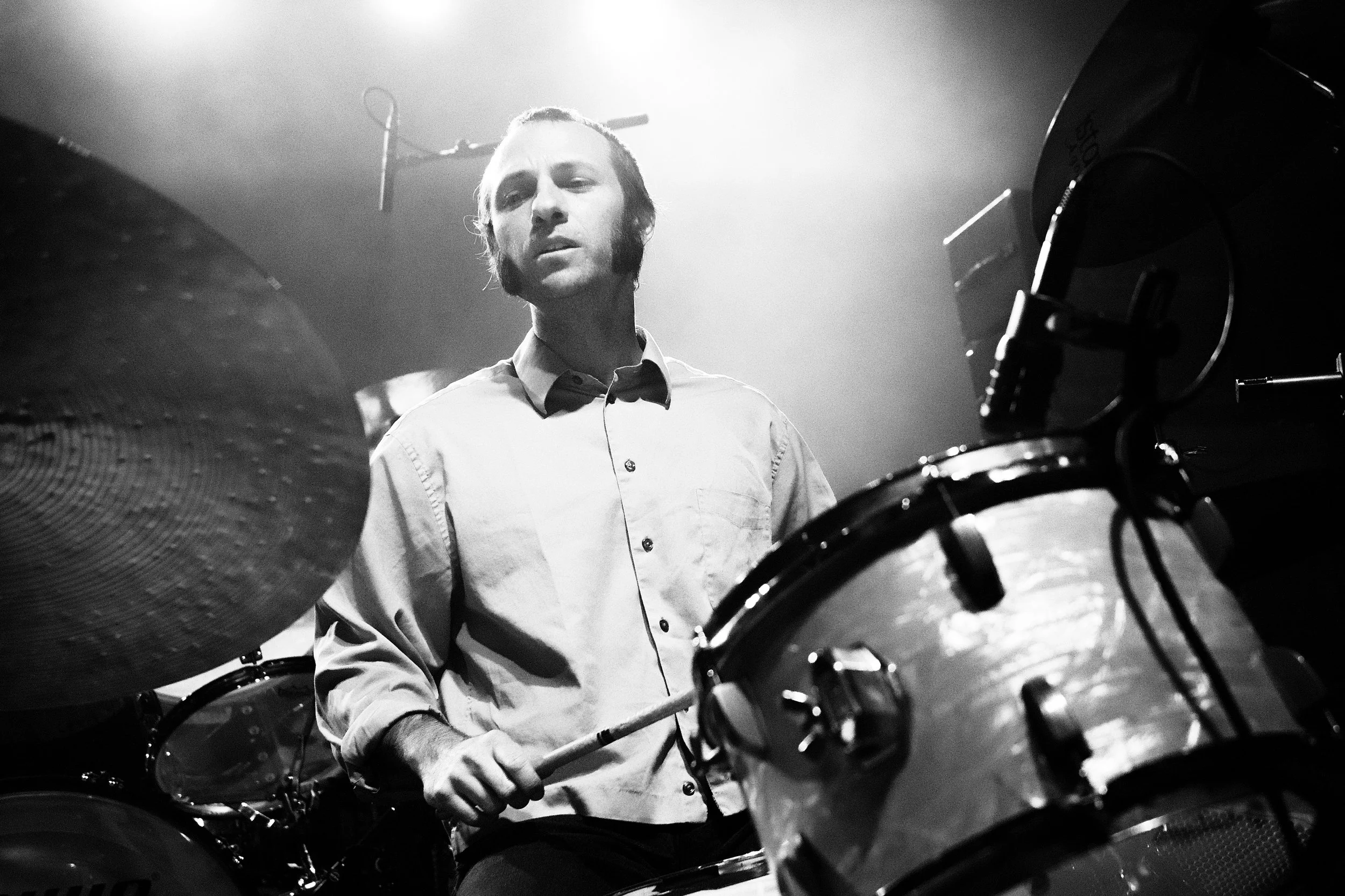 Black and white photo of a male drummer playing drums on stage, with a focused expression, wearing a button-up shirt.