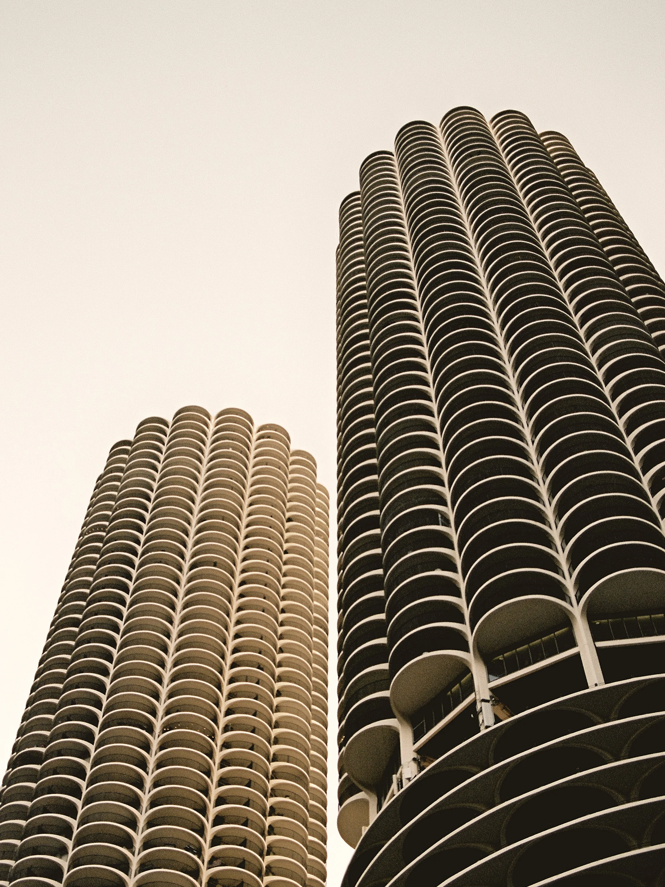 Two tall, modern residential skyscrapers with rounded balconies viewed from below against a pale, cloudy sky.