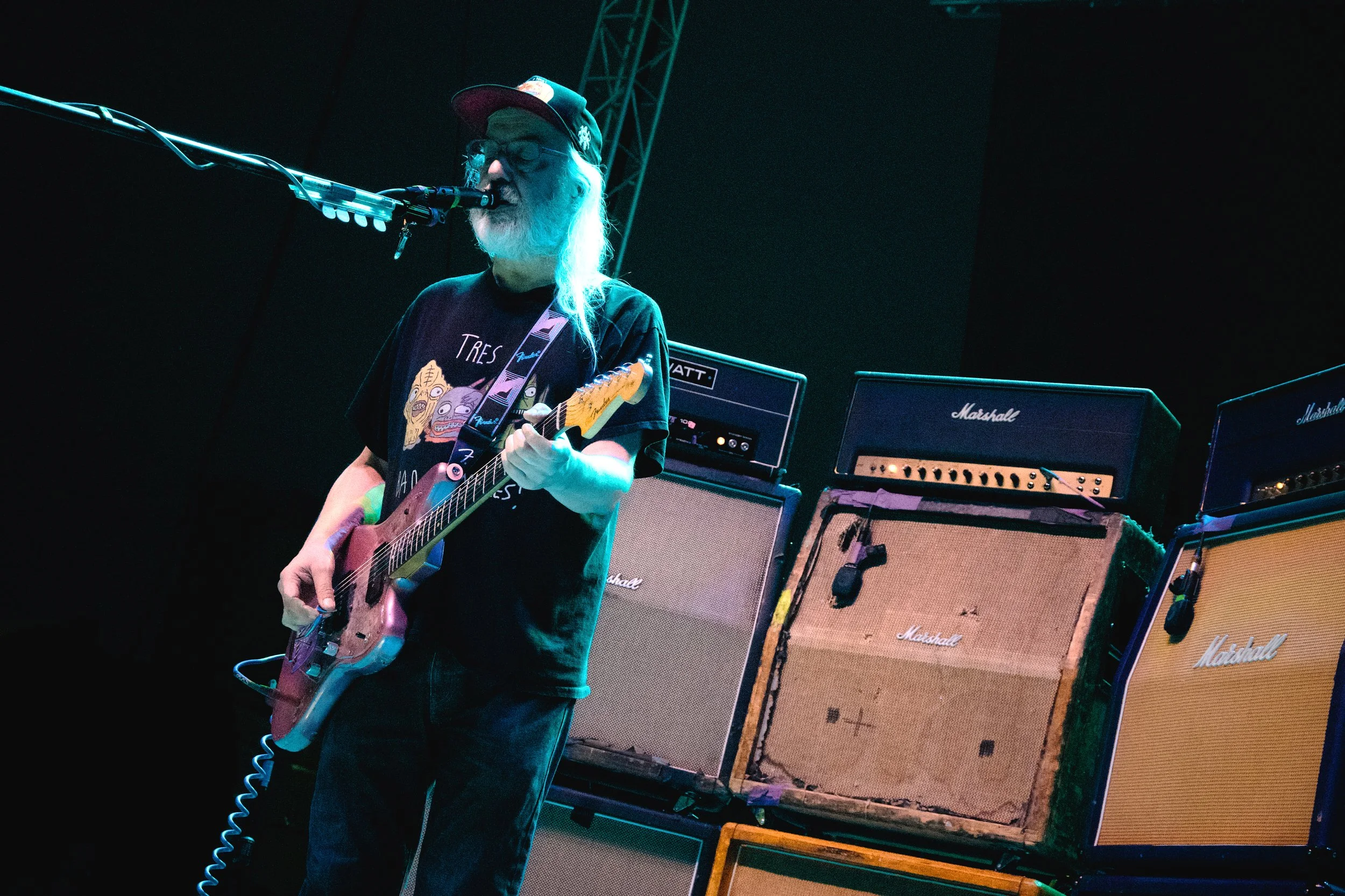 An elderly man with long white hair, beard, glasses, and a black cap is playing an electric guitar and singing into a microphone. He is on stage with several Marshall guitar amplifiers behind him.