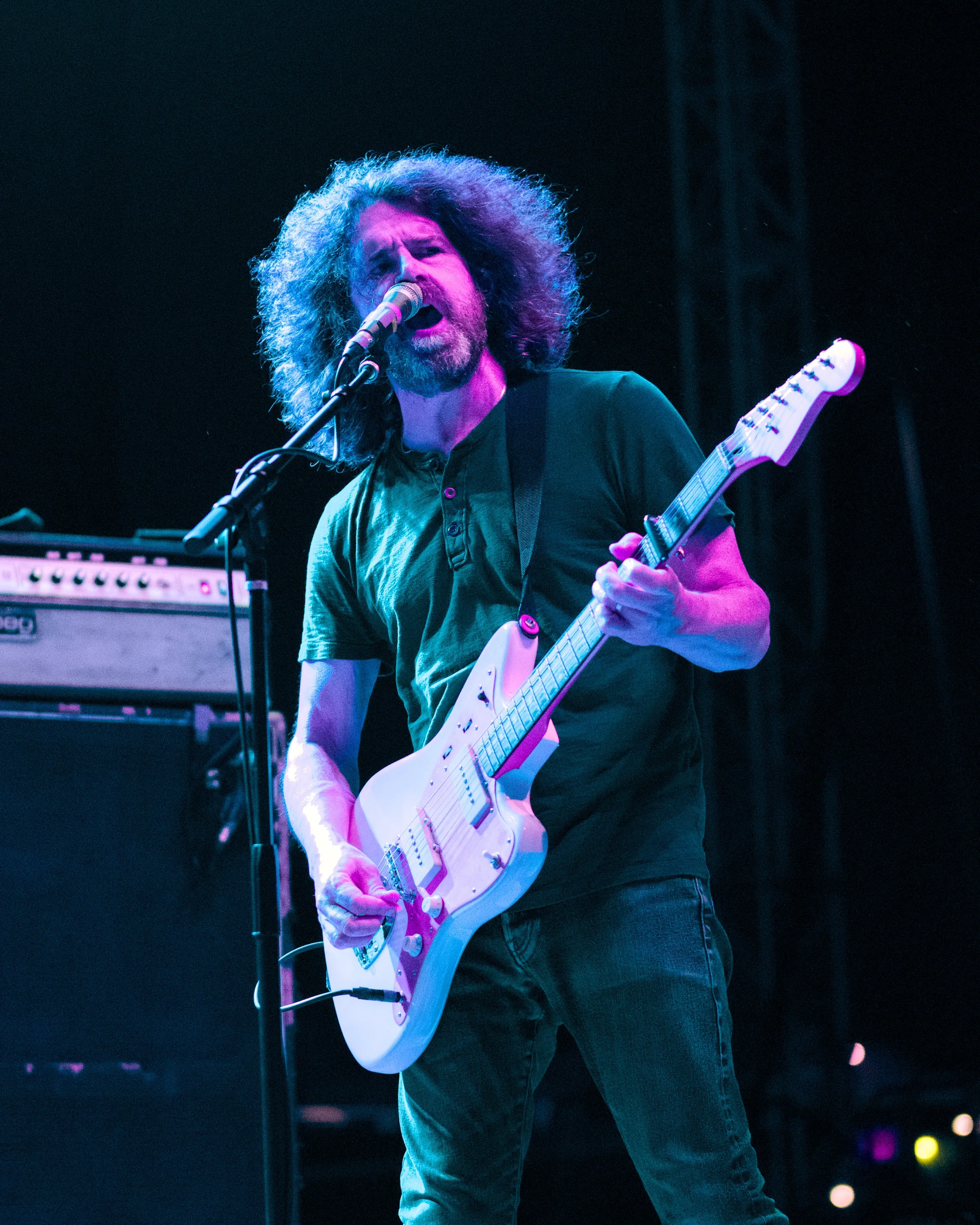 A male guitarist with curly hair singing into a microphone on stage with purple and blue lighting.